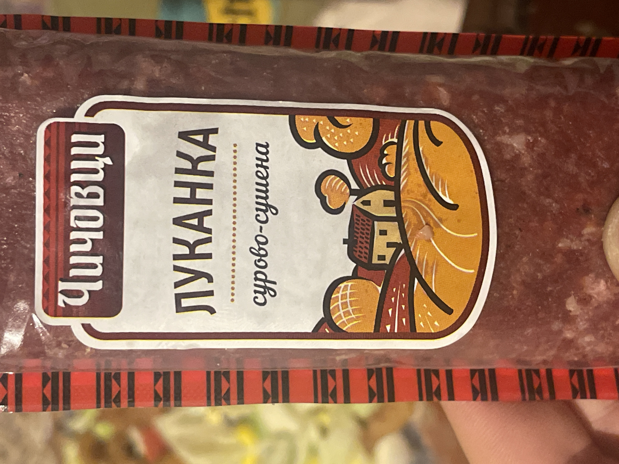 A close-up, slightly angled shot showcases a packaged meat product, likely a sausage or salami, wrapped in clear plastic. The product itself is a dark, reddish-brown color, suggesting a cured or dried meat.
Prominently featured on the package is a white label with a decorative design. On the left side of the label, written vertically in Cyrillic, is "ЧИЧОВЦИ" (Chichovtsi). The central, main text, also in Cyrillic, reads "ЛУКАНКА" (LUKANKA), identifying the product type as a lukanka, a type of dry-cured sausage. Below this, in smaller Cyrillic script, is "сурово-сушена" (surovo-sushena), meaning "raw-dried."
The right side of the label features a stylized illustration in a folk-art aesthetic. It depicts rolling hills and a small, traditional house with a red tiled roof, accompanied by a tree, all rendered in warm tones of orange, yellow, and brown with dark outlines.
The clear plastic packaging has a decorative border at the top and bottom, characterized by a repeating geometric pattern of black shapes on a red background. The background behind the product is out of focus, showing indistinct muted colors, suggesting an indoor setting. No specific activity or event is taking place, beyond the presentation of the food item. The lighting appears to be artificial.
