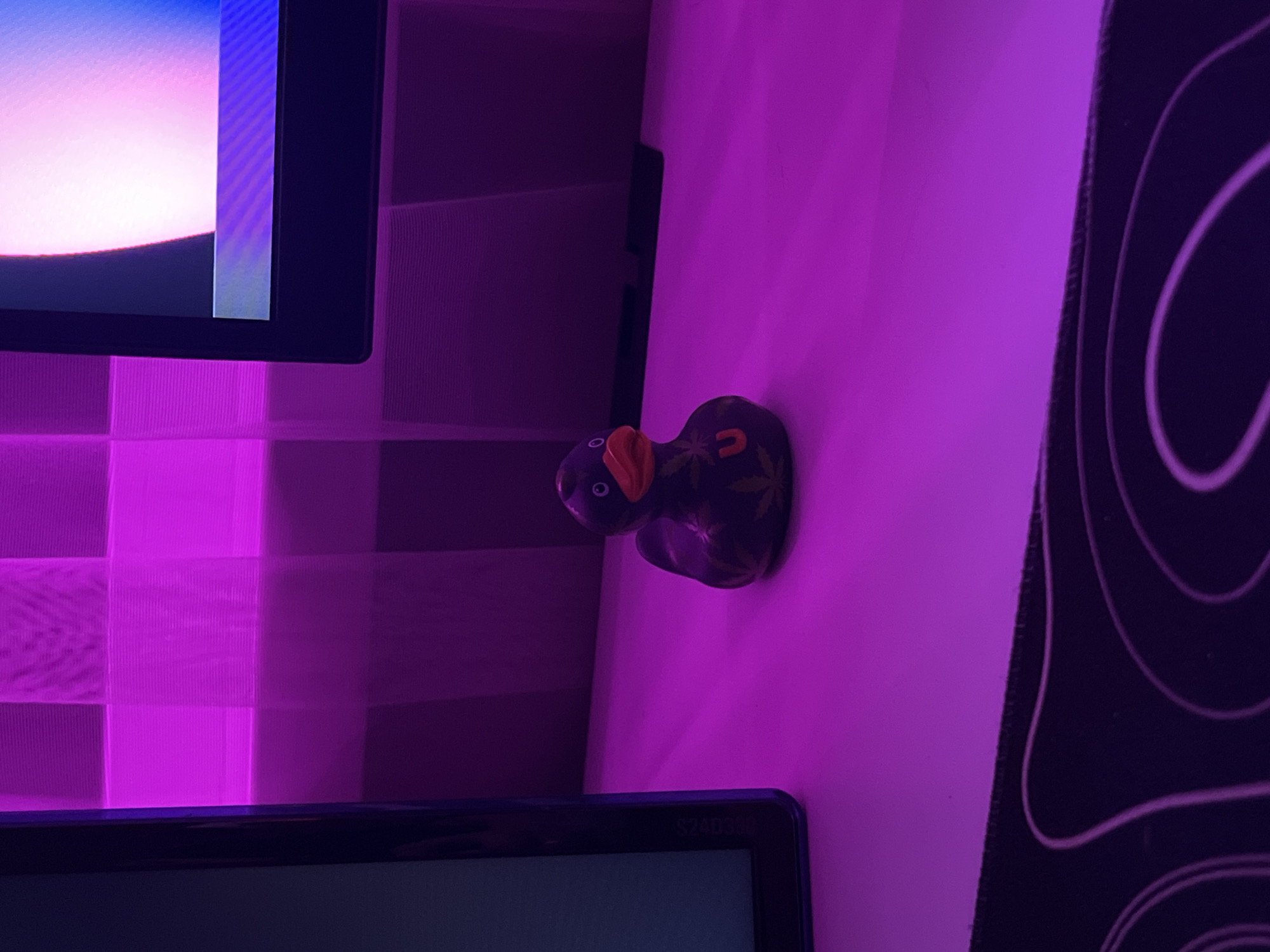 A close-up shot shows a dark purple rubber duck with a red beak and black eyes, sitting on a white surface under purple lighting. The duck has yellow leaf-like patterns on its body and a red 'U' symbol on its chest. In the background, a black monitor is visible, displaying a light pink and blue abstract image. The wall behind the monitor has a checkered pattern illuminated by purple LED strip lights. The foreground features a black mousepad with white wavy lines. The scene is indoors, in an environment that appears to be a desk setup, likely in a bedroom or office.