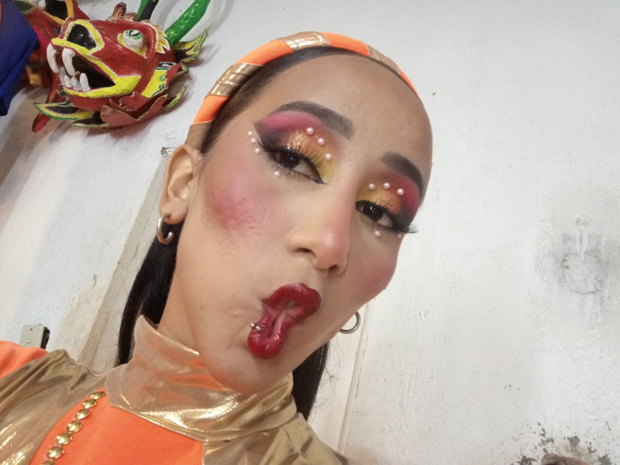 A person with a pierced tongue and heavy stage makeup is posing in a selfie style. The makeup includes pink blush, multi-colored eye shadow with small pearl-like accents, red lipstick, and a matching orange and gold headband. The person is wearing a gold and orange top with gold embellishments. A mask of a dragon-like creature hangs on the white wall in the background. The location is possibly in Porlamar, Venezuela.