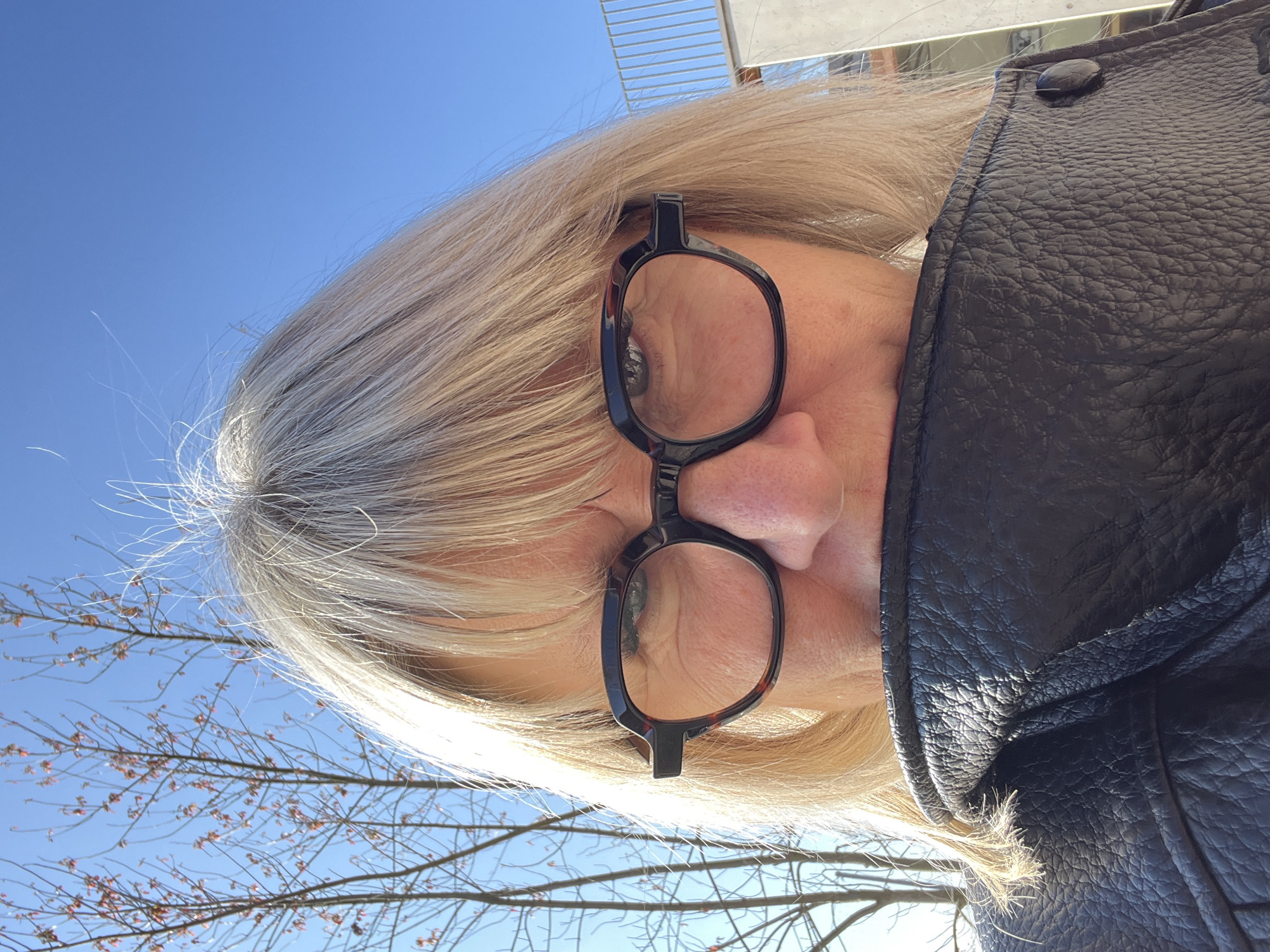 A close-up, slightly angled portrait shows a person with blond, graying hair and wearing black-rimmed eyeglasses. The individual is also wearing a black leather jacket. Behind them, a clear blue sky is visible, along with bare tree branches and part of a building with a metallic railing. The location is La Fouillouse, France.