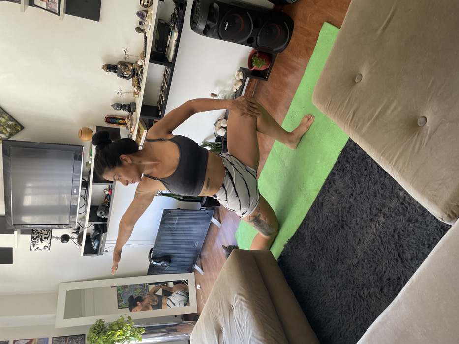 This image depicts a woman performing a yoga pose within a brightly lit indoor setting, likely a living room in Buenos Aires, Argentina. She is positioned on a vibrant green yoga mat, with her left knee down and her right foot flat on the mat, her body in a twisted lunge. Her right hand rests on her right knee, while her left arm is extended outwards to the side. She is wearing a grey sports bra and white and black striped shorts, with her dark hair pulled back in a bun.
The background reveals a home environment, with a large television screen mounted on a wall. Below the TV and extending to the right are shelves adorned with various decorative objects, including Buddha statues, crystals, and small potted plants. A large black speaker system is visible on the floor to the right. A light-colored sofa or cushion is partially visible to the right, adjacent to a dark grey shaggy rug that borders the yoga mat. A large white-framed mirror on the left wall reflects the woman in her yoga pose, and a green plant is placed in front of it. The lighting suggests it is daytime. There is no visible text legible within the frame.