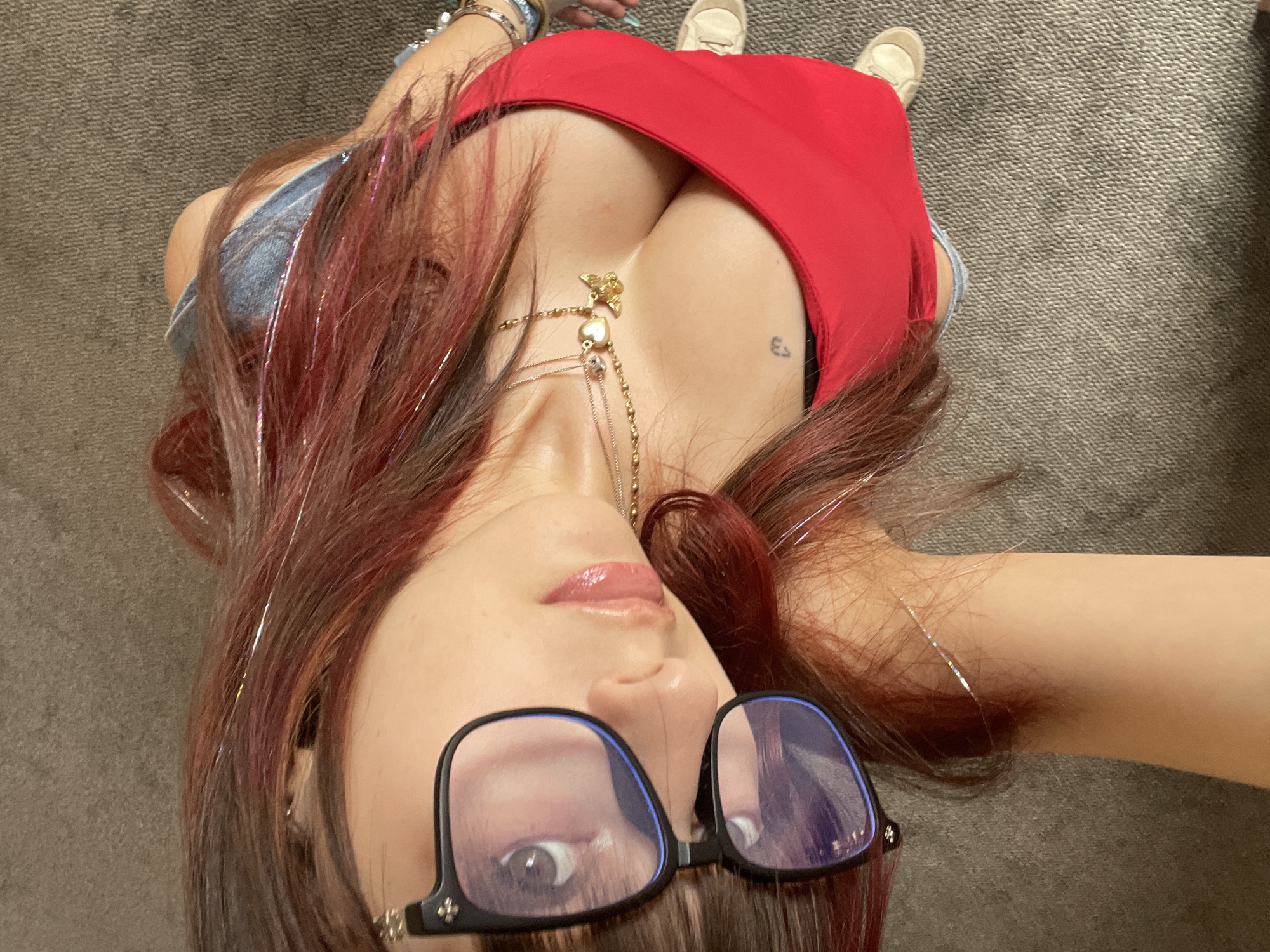 The image shows a close-up, overhead view of a person lying down. The person has long, reddish-brown hair with some sparkly strands. They are wearing large black-rimmed glasses with blue-tinted lenses, a red tank top, and denim shorts. Two necklaces are visible, one with a heart charm and another with a butterfly charm. A small, dark tattoo is visible on the chest. The person's skin is pale, and they have glossy, pink lipstick on. Their right arm is bent, with the hand near their face. The background is a textured, grey surface, likely carpet or upholstery. The lighting is warm and somewhat dim. There is no indication of a specific location in London, United Kingdom, or any other city. The scene appears to be indoors, in a casual setting.
