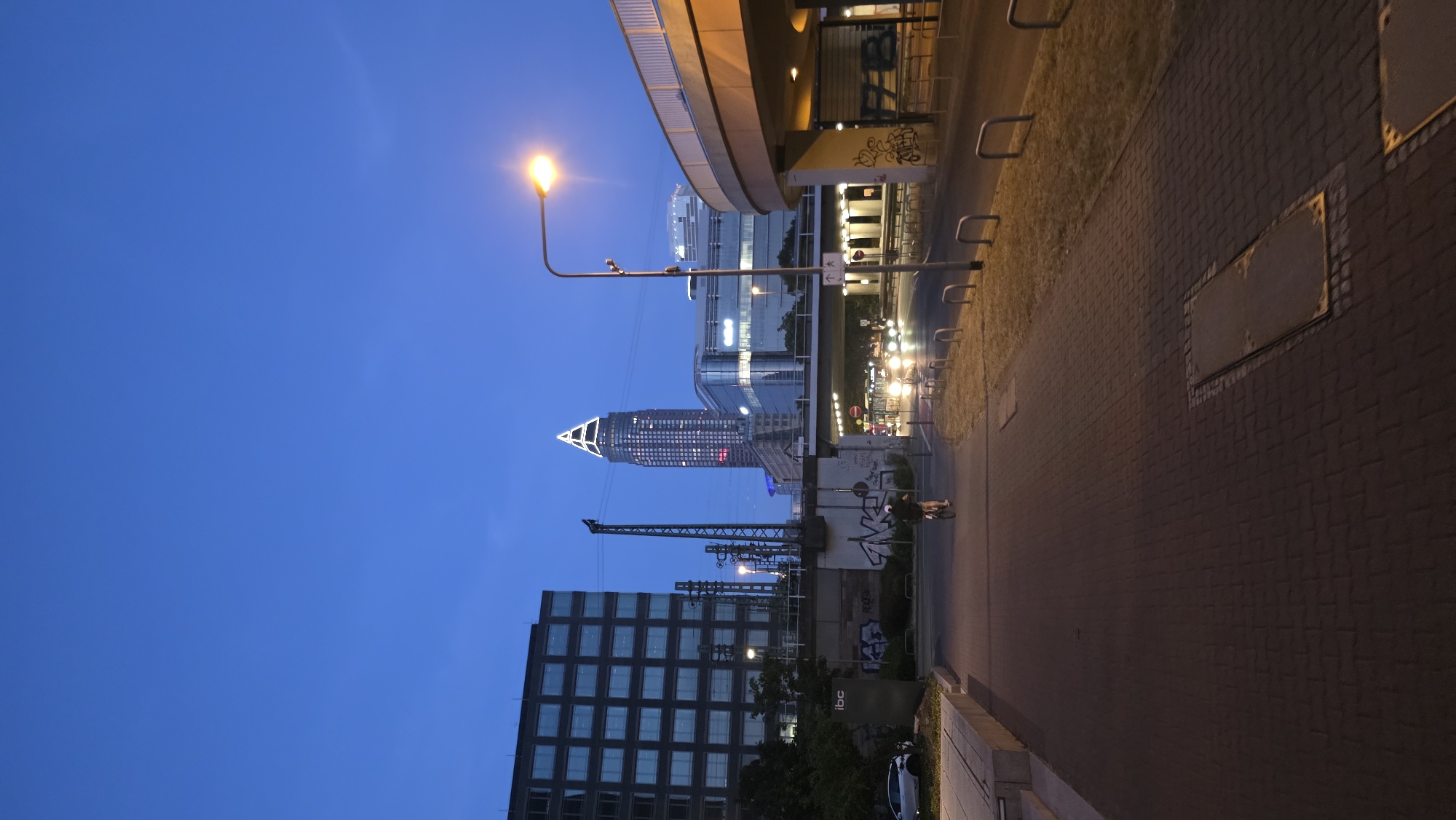 The image depicts an urban scene at dusk in an unknown city. In the foreground, a paved walkway runs towards the viewer, bordered by a grassy area with U-shaped metal bollards on the right. A person is cycling away from the camera on the walkway.
In the midground, a road with oncoming headlights is visible under an elevated bridge. To the left of the walkway, a dark, multi-story building with a grid of windows stands prominently. Behind this, a tall, triangular-shaped skyscraper, illuminated with white light at its peak, dominates the skyline. Several other modern buildings with glass facades are also visible.
The sky is a deep blue, indicating twilight. A streetlamp with a glowing orange light is positioned on the right side of the image, illuminating the immediate surroundings. Graffiti is present on a concrete wall beneath the elevated bridge. A sign that reads "ibc" is visible on a post to the left. The overall impression is a bustling urban environment transitioning into night.