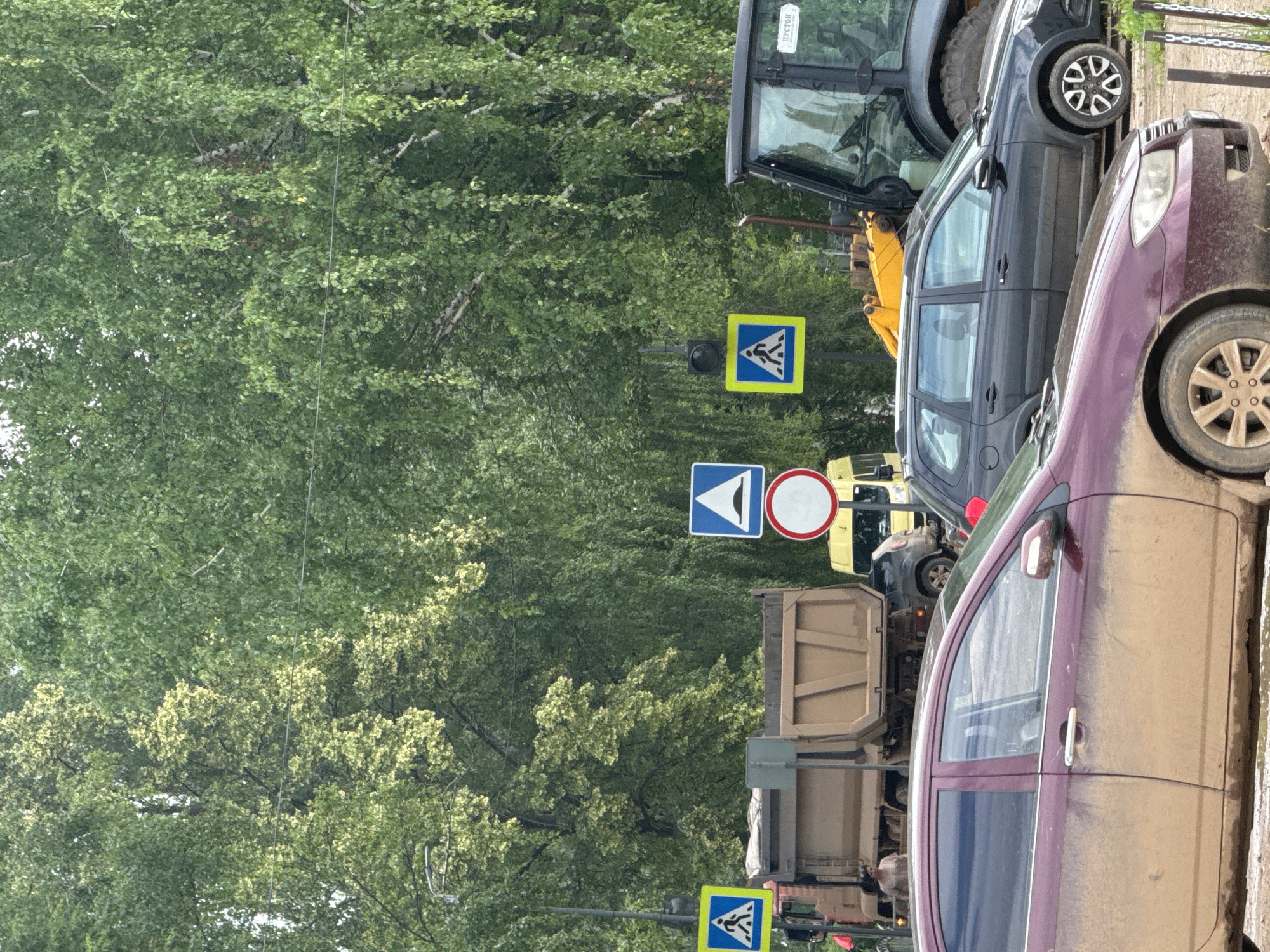 In Chastyye, Russia, a scene unfolds on a street lined with lush green trees. Several vehicles are present, including a burgundy car in the foreground, appearing muddy, and a dark-colored station wagon parked behind it. Further back, a large beige dump truck is visible, and a black construction vehicle with a yellow loader is to the right. Traffic signs are scattered throughout the image, including pedestrian crossing signs and a sign indicating a speed bump. The overall impression is that of an urban street during the daytime, with ongoing construction or road work suggested by the presence of the construction vehicle and the muddy conditions.