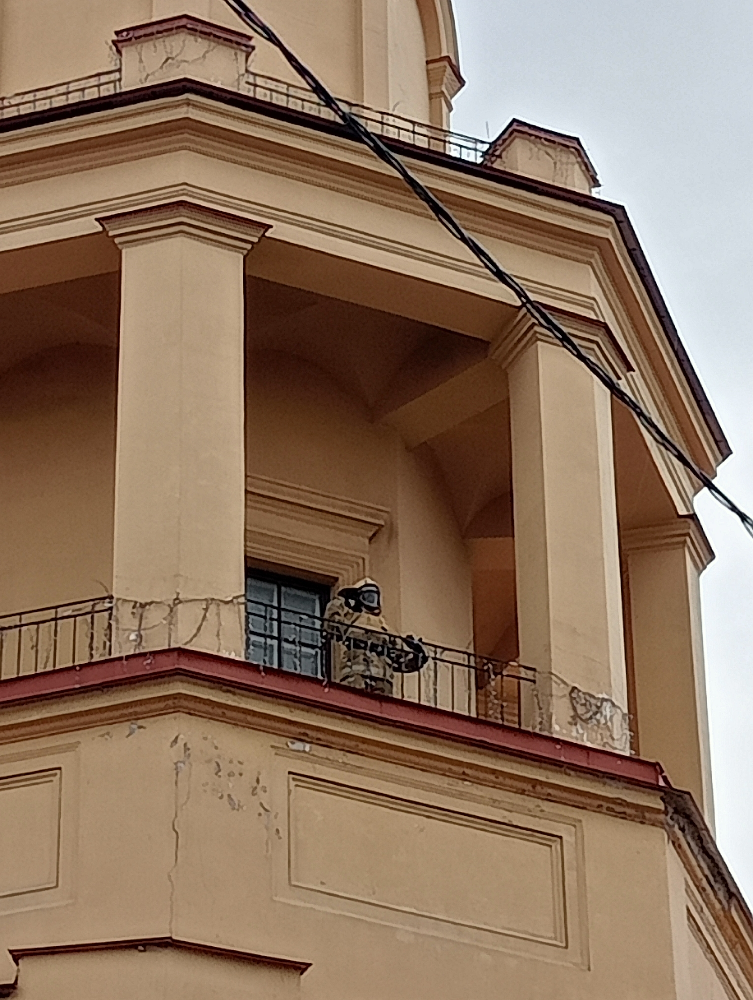 The image shows a person in a hazmat suit standing on a balcony of a building. The building appears to be made of light-colored stone and has a red roof. There are columns and arches visible, suggesting it is an older, possibly historical, structure. The hazmat suit is white with dark accents and the person is wearing a full-face respirator. They are holding what appears to be equipment or tools. A window is visible behind the person. Electrical wires are strung across the upper portion of the image. The sky is overcast. Based on the architectural style and the overall aesthetic, this building is likely located in Saint Petersburg, Russia.