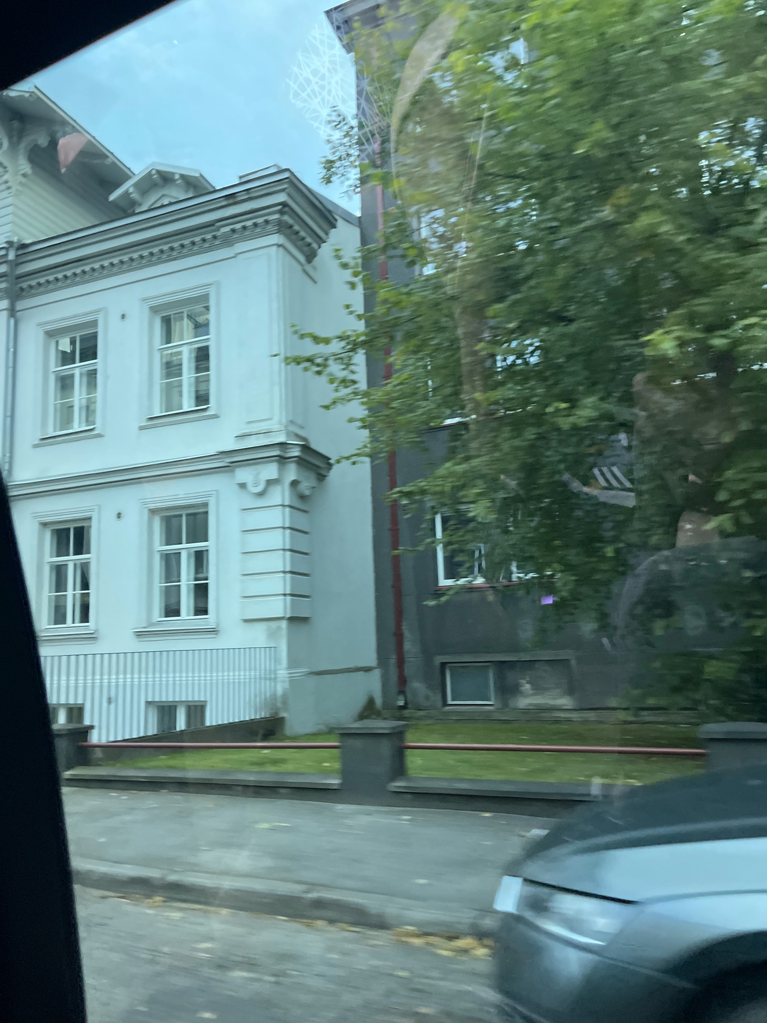 This image captures a view of buildings and trees, possibly from a moving vehicle in Tallinn, Estonia. The left side of the frame features a pale blue, multi-story building with white-trimmed windows, decorative cornices, and a striped awning. Adjacent to it, on the right, is a darker-colored building partially obscured by lush green trees. A grey car is visible in the foreground, its headlights and hood catching the light. A sidewalk and a grassy area with a red railing separate the street from the buildings. The overall lighting suggests it is daytime, with the sky visible through reflections on the glass.