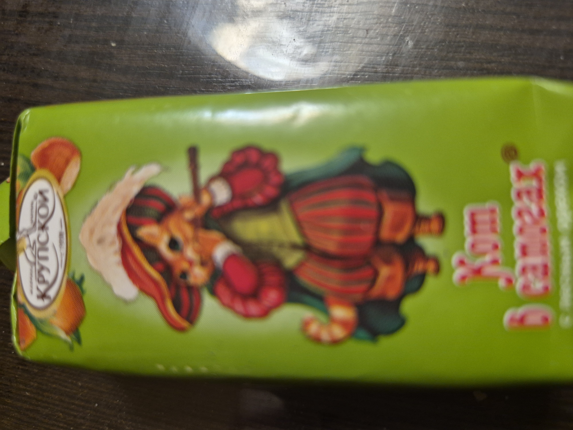 The image shows a green wrapper with a cartoon illustration of a cat wearing a hat and boots. The brand name "КРУПСКОЙ" is visible on the left side of the wrapper. There is also some text in Russian language on the right side, "Кот в сапогах." The wrapper appears to be lying on a dark wooden surface. The location is unknown.
