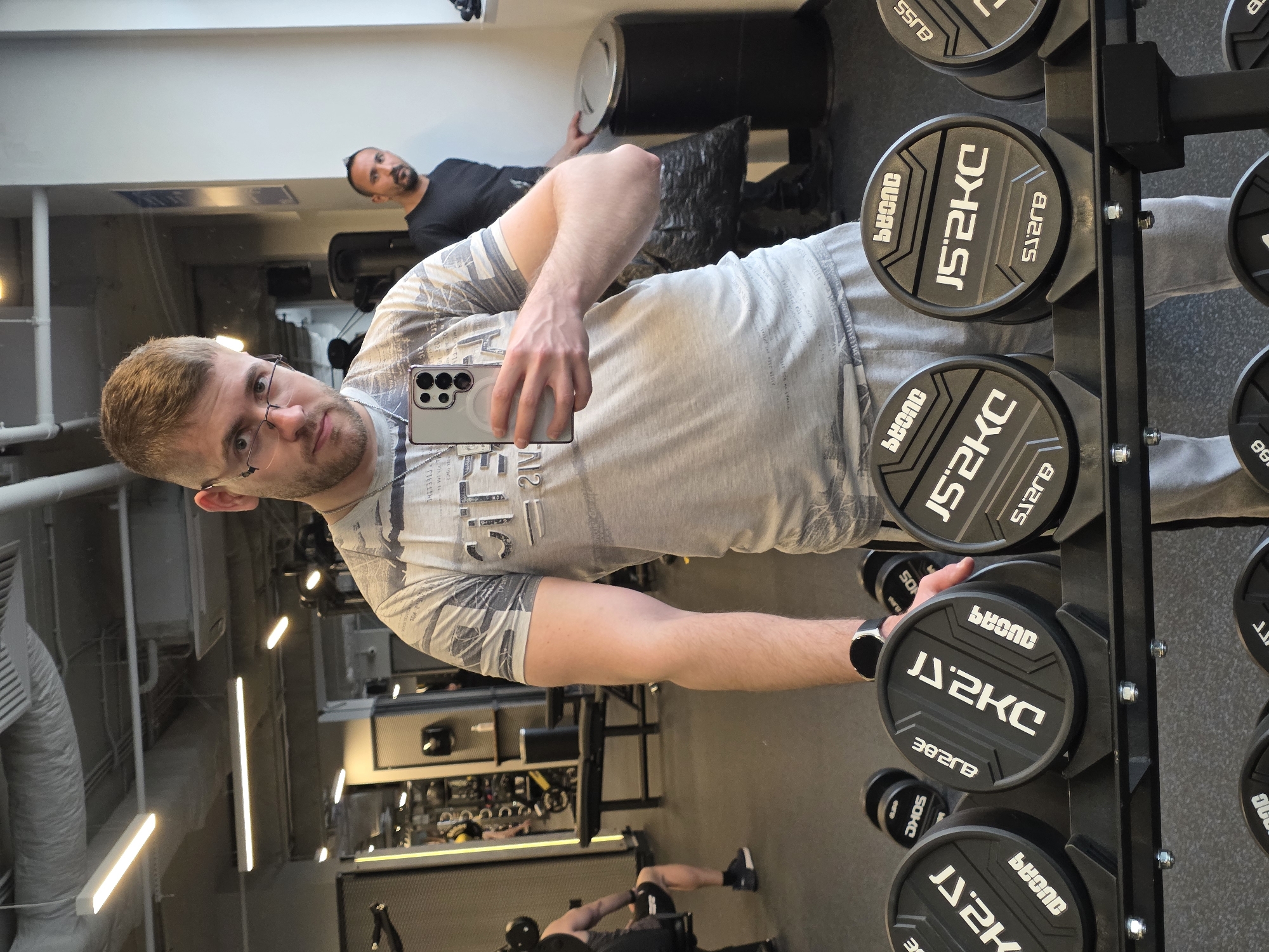 The image shows a man in a gym taking a selfie in front of a rack of 2.5 kg weights labeled "JS2KC". He is wearing glasses, a light-colored t-shirt with writing on it, and a smartwatch. There are other people visible in the background of the gym, including a man lying on a bench and another person working out on the floor. The gym appears to be well-lit with fluorescent lighting and has various workout machines and equipment. The location cannot be determined to be Plovdiv, Bulgaria from this image.