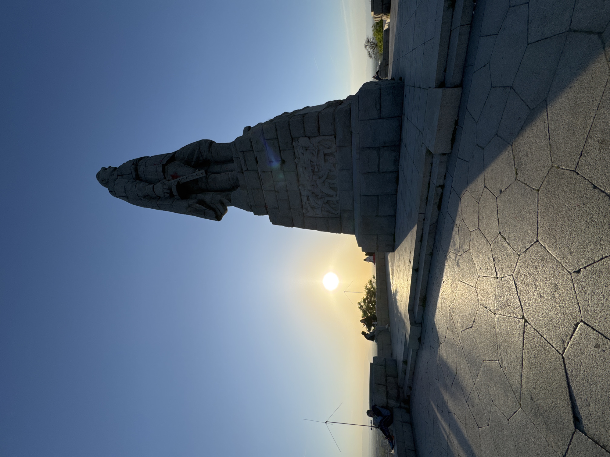 The image captures the Alyosha Monument in Plovdiv, Bulgaria, at sunset. A towering stone statue of a Soviet soldier, holding a rifle and wearing a helmet with a faint red star detail, dominates the left side of the frame, silhouetted against the bright sky. The monument's base features carved reliefs depicting figures.
The scene unfolds on an elevated, paved viewing platform, constructed with large, irregularly shaped hexagonal stone slabs. The late afternoon or early evening sun, a brilliant white disc, hangs low in the sky, casting long shadows across the ground and highlighting the monument's form. The sky is clear, transitioning from deep blue overhead to a warm, golden hue near the horizon.
Several people are visible on the platform. Some are seated or standing near the monument's base and along a low wall, appearing to observe the sunset or the distant landscape. One person in the foreground is seated, with what appears to be an antenna-like structure nearby. The overall atmosphere is serene and contemplative, enhanced by the golden hour light. No discernible text is visible on the monument or in the surrounding area.