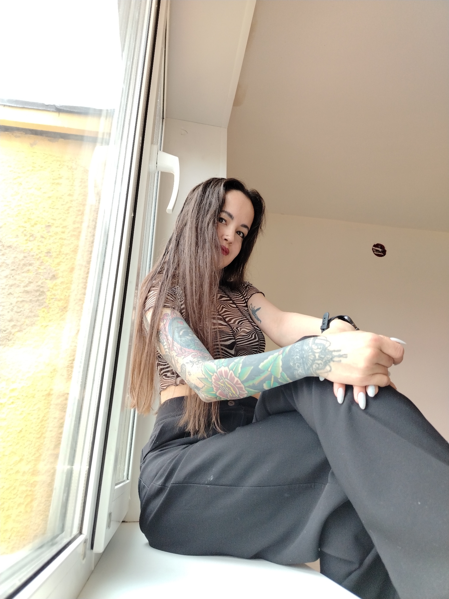 A young woman with long, dark brown hair is seated on a white window sill in Noviy, Russia, looking directly at the camera. She is wearing a short-sleeved, brown and black striped crop top and black trousers. Both of her forearms are extensively covered in detailed, colorful tattoos, including floral patterns. She has red lipstick, a small nose piercing, and long, light-colored manicured nails. A dark wristband or watch is visible on her left wrist.
The setting is indoors, next to a large, white UPVC window that allows bright daylight to illuminate the scene. Through the window, a textured, yellow exterior wall is visible. The interior wall behind the woman is a plain, light beige, and there is a small, circular cutout or hole in the wall, possibly an unfinished electrical fixture. The photo is taken from a low angle, looking up at the subject. The time of day appears to be daytime, with ample natural light. No visible text is present.