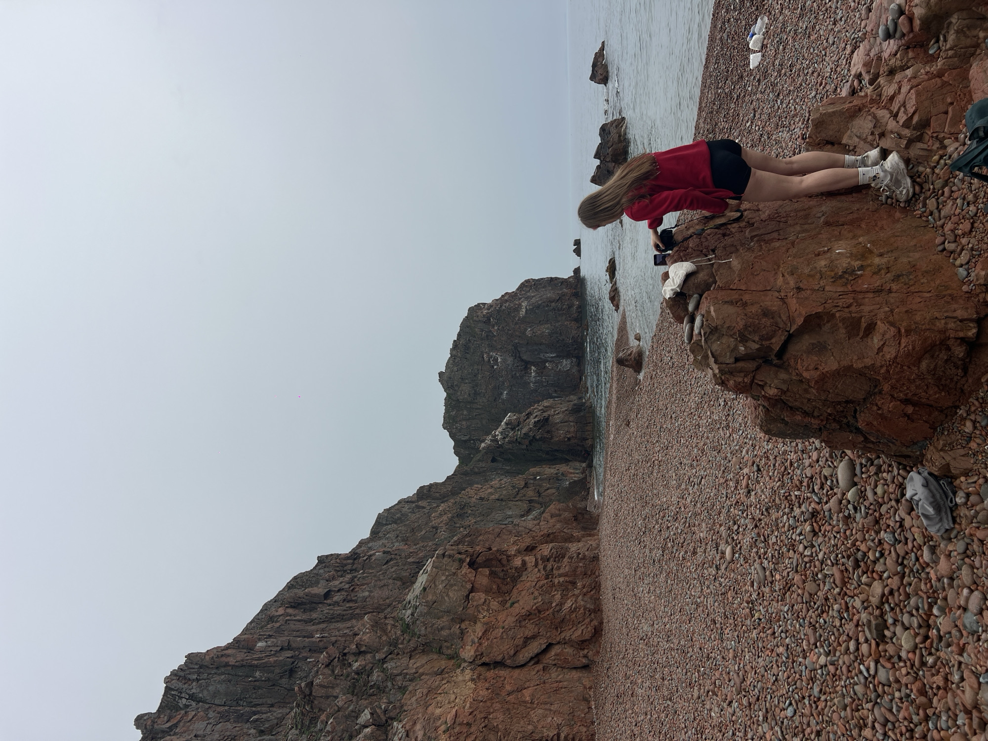 A woman in a red sweatshirt and black shorts kneels on a large rock at a beach on Russkiy Island, Russia, holding a camera and pointing it towards the ocean. She is wearing white sneakers and white socks. The beach is covered in small reddish-brown stones. Behind the woman are other rocks and the ocean. The sky is overcast.
