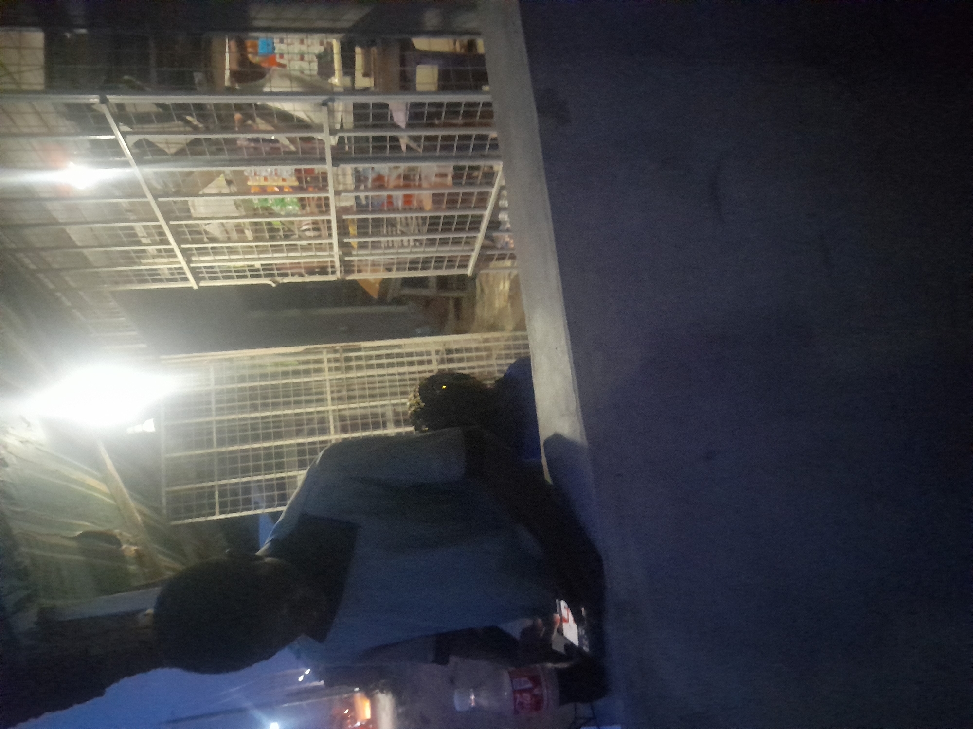 This nighttime image, captured in Ogbomosho, Nigeria, shows an indoor or semi-outdoor retail space, likely a shop or stall.
A dark-skinned individual, wearing a light-colored short-sleeved shirt, is positioned in the left foreground. They appear to be standing or leaning forward, looking down at something on a surface. Part of another person's head and hair is dimly visible behind them.
The background is dominated by multiple white, wire-frame shelving units, characteristic of a small store. These shelves are stocked with various items, including what appear to be bottled drinks and packaged goods, though specific details are obscured by distance and lighting. A prominent, bright artificial light source is visible on the upper left, casting a strong glare and creating uneven illumination throughout the scene. A thick, light-colored pillar or wall runs vertically along the right side of the frame.
On a surface in front of the main person, a white plastic bottle with a distinct red label is visible. The text "Coca-Cola" is discernible on this label. The overall ambiance is dark, with pockets of bright light, confirming the nighttime setting. The activity of the main person is unclear, but they seem to be focused on an object or task directly in front of them.