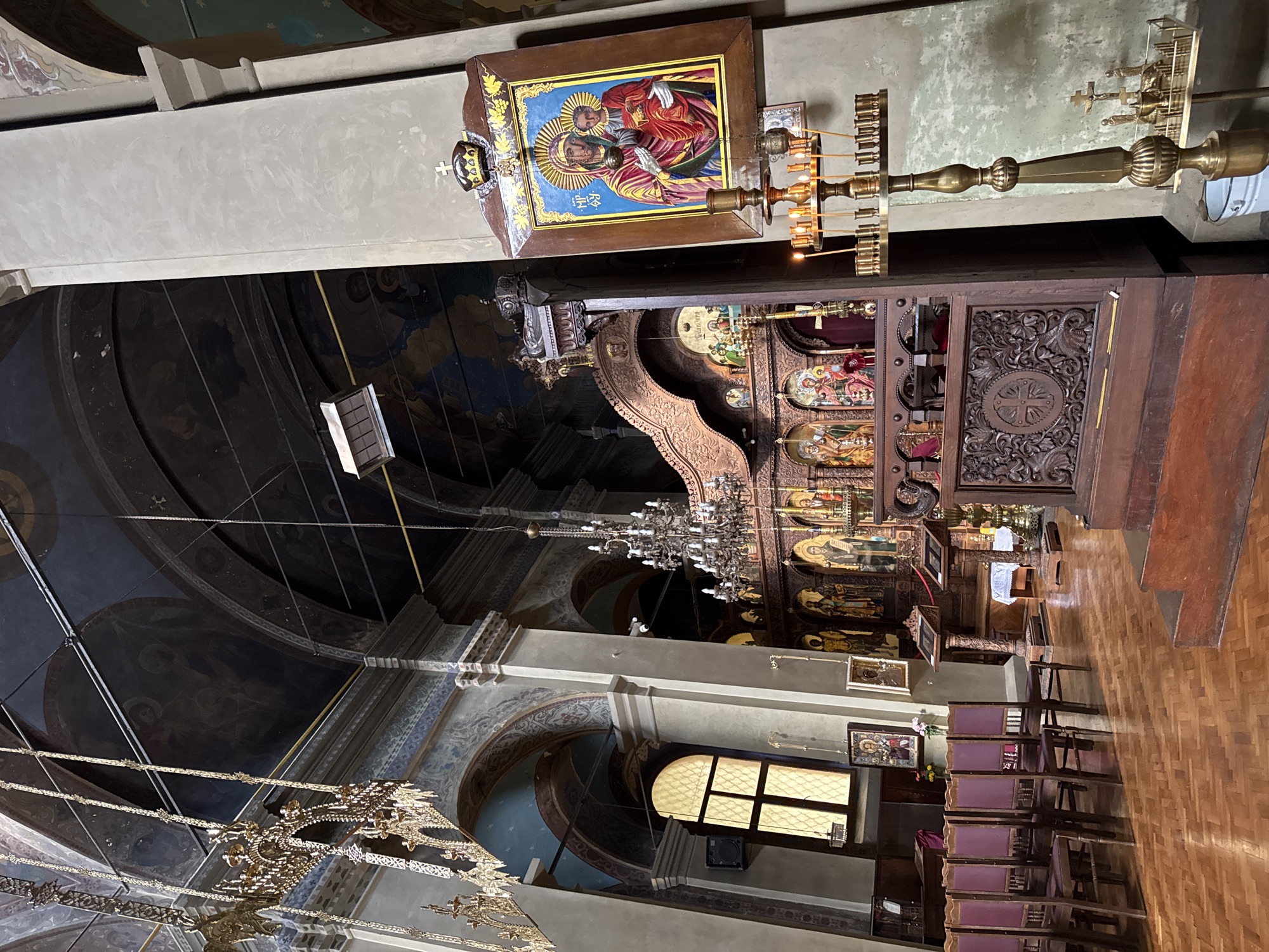 The image shows the interior of a church. In the foreground, there is a wooden iconostasis, adorned with various religious icons. To the right, a tall brass candelabrum with lit candles stands prominently. A large icon of the Virgin Mary and Child hangs on the wall to the right of the candelabrum. In the background, the church ceiling is decorated with frescoes. Rows of wooden chairs with red upholstery are visible on the left side of the image. The floor is made of polished parquet. This is an interior view of the Saint George Church in Yambol, Bulgaria.