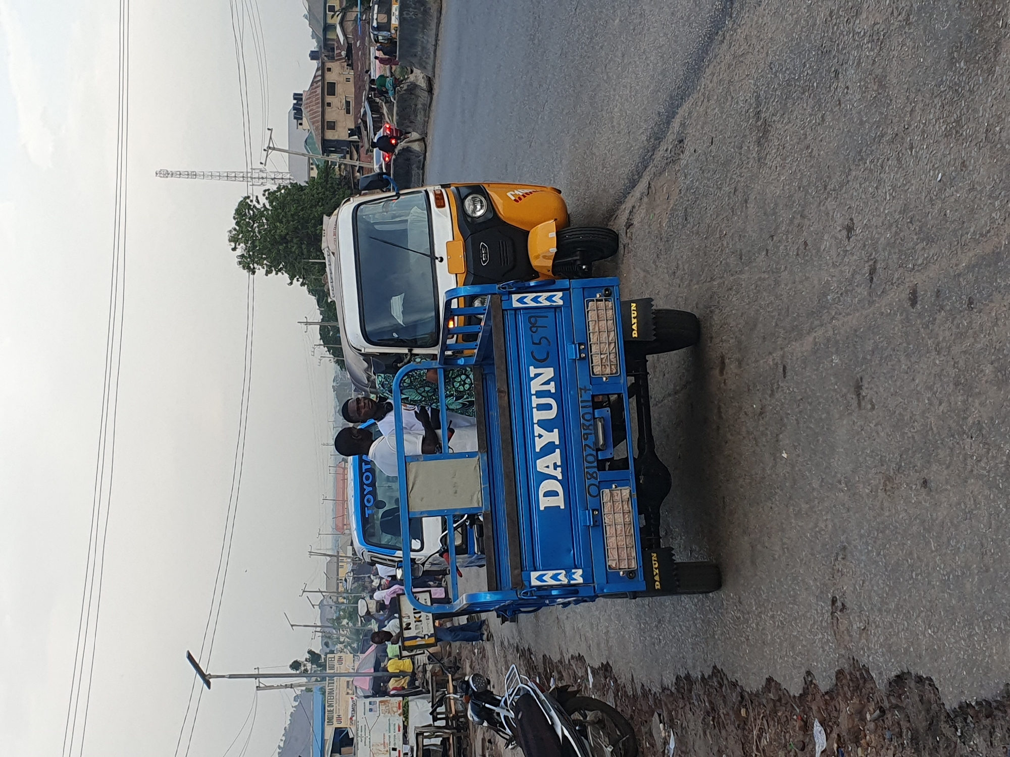 The image depicts a blue and yellow three-wheeled vehicle with "DAYUN" printed on the side, partially obstructing a street in Lokoja, Nigeria. Two passengers are visible inside the vehicle. The road is paved and bordered by a dirt shoulder with trash and debris. Buildings and overhead power lines are present in the background, along with people gathered on the side of the road near a sign. A motorcycle is parked in the foreground.