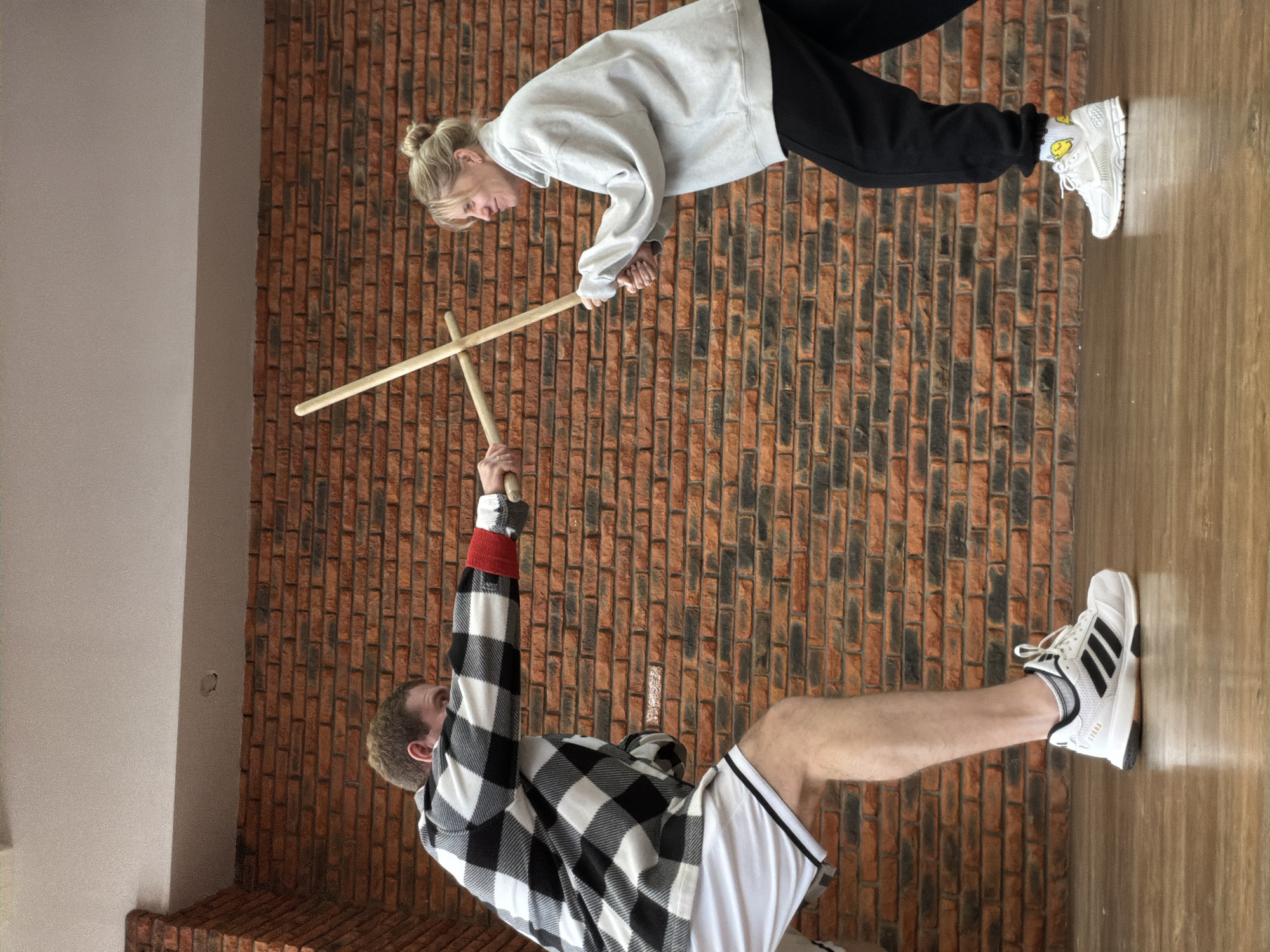 Two people are engaged in what appears to be a martial arts practice with wooden sticks in an indoor setting. A woman with blonde hair in a bun, wearing a light grey sweatshirt and black pants, is holding one stick horizontally. Facing her is a man in a black and white checkered shirt and white shorts, who is holding the other stick vertically. The background is a brick wall and a light-colored wooden floor. Based on the wall design and architectural style, it is likely that the location is in Chaikovsky, Russia.