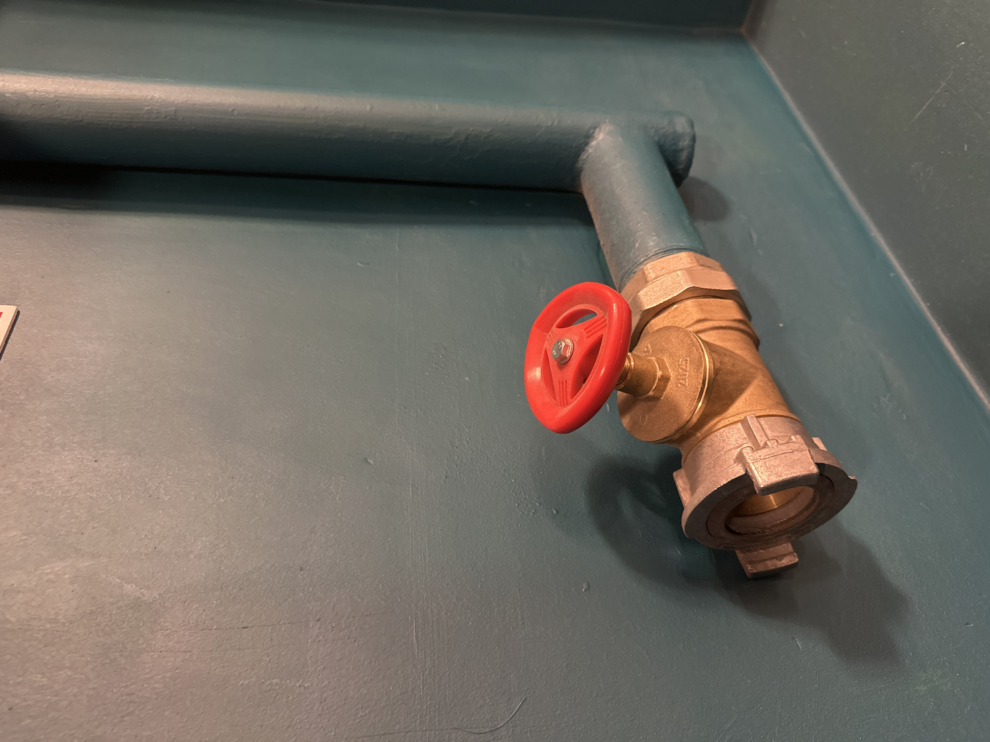 A close-up shot presents a metal pipe affixed to a wall, featuring a prominent valve with a red handle. The scene lacks any human presence, indicating a static, inanimate environment. The wall and pipe are painted in a similar shade of teal or light blue, creating a monochromatic backdrop that highlights the red valve handle. The valve's brass components contrast with the painted surfaces, suggesting a functional element within a building's infrastructure. There is no information to confirm that the location is in Moscow, Russia.