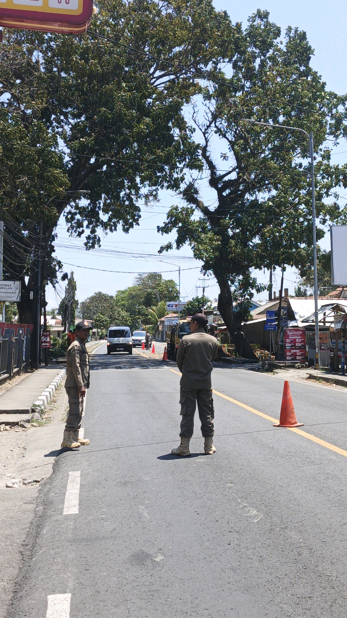 In the city of Sukaresik, Indonesia, two individuals in matching tan uniforms and caps stand on the asphalt of a road. The person on the left faces the camera, while the person on the right faces away. Both are wearing tactical-style pants and tan boots. The road has a yellow line marking the center. Orange traffic cones are placed on the road, suggesting a temporary closure or checkpoint. In the background, trees with dense green foliage arch over the road, forming a natural canopy. A white van and a car are visible further down the road. Buildings and stalls line the right side of the street, indicating an urban or semi-urban environment. Power lines and street lights are present. The sky is clear and blue.