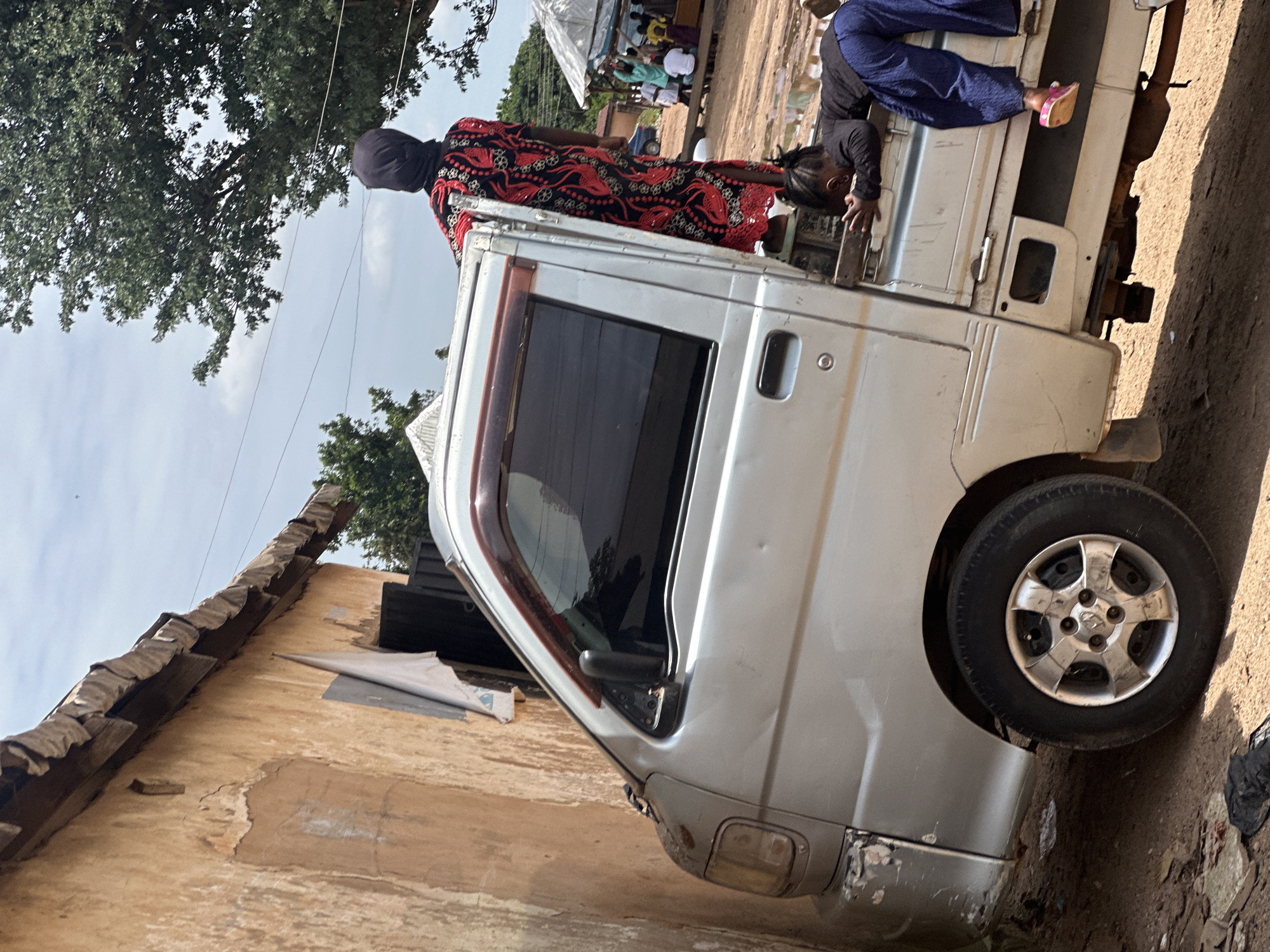 In Barakin Lerawi, Nigeria, a silver pickup truck is parked precariously close to a damaged, ochre-colored building. The truck's driver's side is pressed against the building's rough-textured wall, with a white cloth hanging from an opening near the top. A person wearing a black headscarf and a red and black patterned dress stands on the edge of the truck bed, their hands on the roof. In the truck bed, a young child with braided hair, dressed in a black long-sleeved shirt and blue pants, is lying face down, resting their arms on the truck's edge. Their legs are bent, and one pink sandal is visible. In the background, a large green tree provides shade, and beyond it, more trees and a cloudy sky are visible. Further in the background, several people are gathered under makeshift tents, with a blue car parked nearby. The ground surrounding the truck is a mixture of dirt and debris, with evidence of recent rain or water puddles.