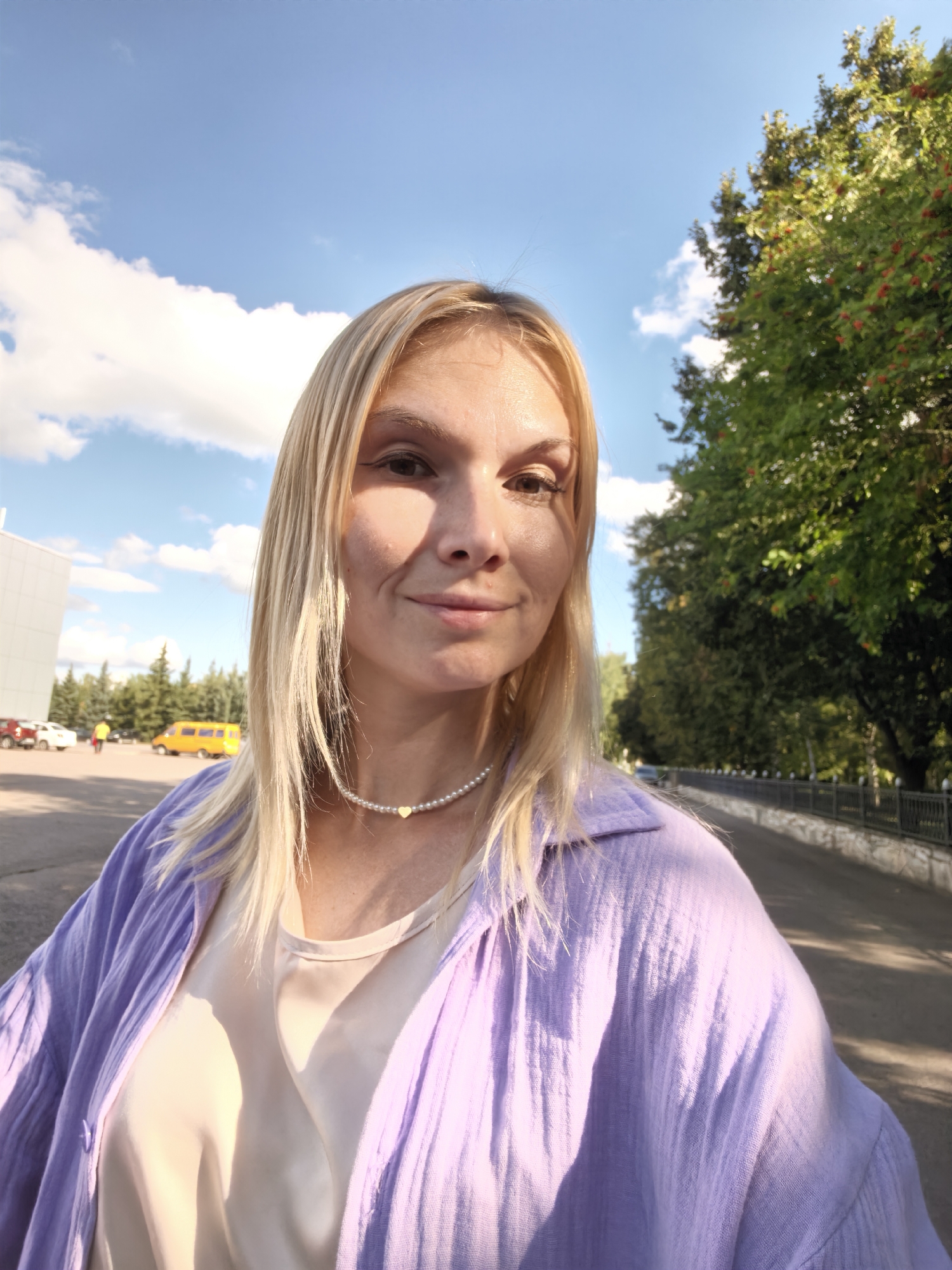 The image is a medium shot selfie of a blonde woman in Sterlitamak, Russia. She is standing outdoors, with a blue sky and scattered clouds above. To her right is a lush green tree with red berries, and to her left, a modern building and a parking lot with several cars and a yellow van. She is wearing a light-colored blouse under an unbuttoned, loose-fitting purple shirt. A pearl necklace with a small heart pendant is visible around her neck. The woman is looking at the camera with a slight smile. The overall setting appears to be a public area, possibly a school or university campus, given the presence of the building and the van that resembles a school bus. The lighting suggests it is daytime, likely late afternoon, with the sun casting shadows.