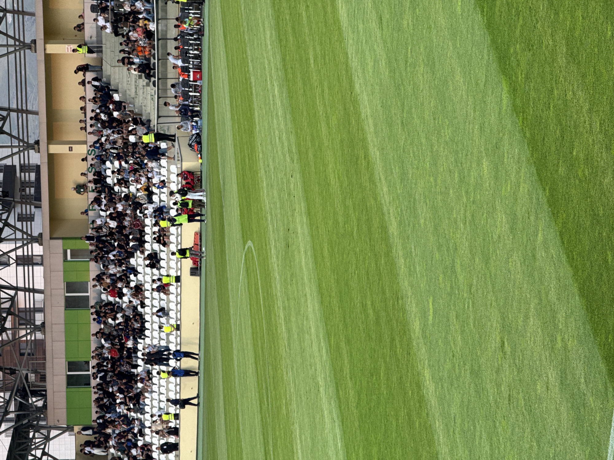 The image shows a portion of a football stadium with spectators seated in the stands. The stands are filled with people, some of whom are wearing casual clothing, while others appear to be staff members in high-visibility vests. The field, covered in meticulously maintained green grass with visible white markings, occupies the majority of the frame. The angle of the shot suggests it was taken from an elevated position, possibly from another part of the stadium or an adjacent building. The lighting indicates it's daytime, and the scene appears to be outdoors. The focus is on the convergence of the crowd and the playing field, capturing a moment during a sporting event. The location is identified as Abovyan, Armenia. There are no overt emotional expressions visible due to the distance and resolution, but the presence of spectators implies a game or a related event is taking place. The overall atmosphere seems to be that of a typical stadium environment during a sporting occasion. Visible text includes "MACRON" on a sports jersey.