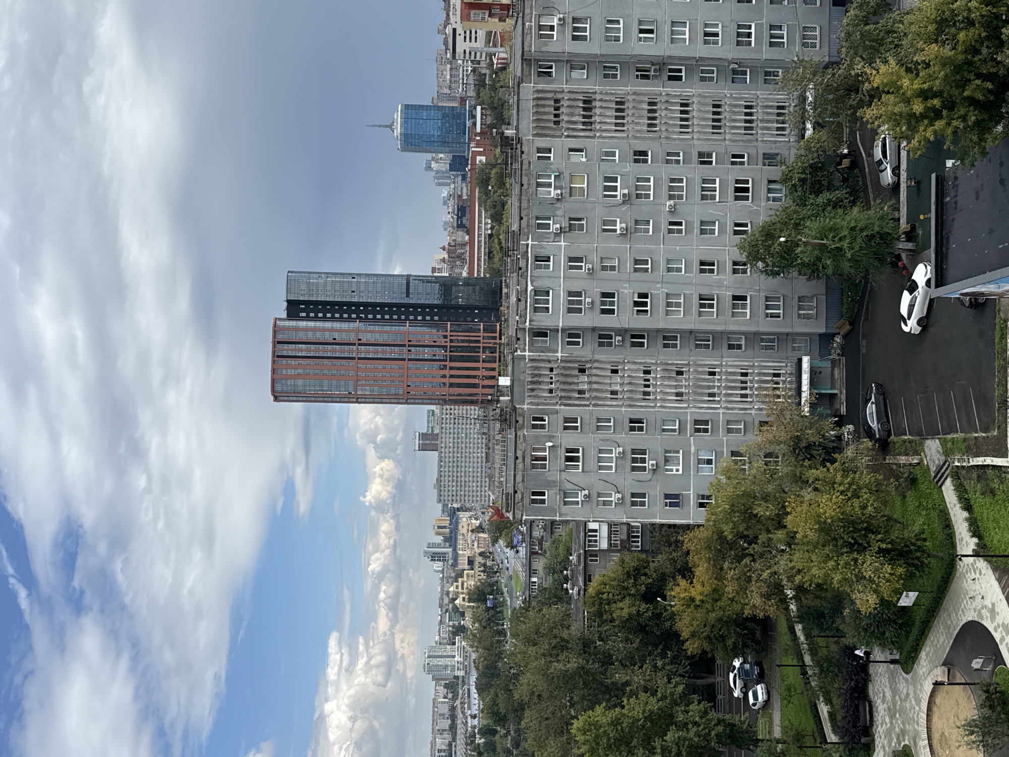The image captures a cityscape with a focus on residential and commercial buildings under a cloudy sky. In the foreground, a tall, grey, apartment building dominates the lower half of the frame, its facade characterized by numerous windows and visible air conditioning units. To the right of the apartment building, a darker, narrower building is visible. Further in the background, a prominent red and black skyscraper stands out, with two sleek, modern high-rises to its right, one with a distinctive blue, rounded glass facade and a spire. The middle ground features a street with cars and trees lining its sides, and a variety of other buildings, some appearing to be of moderate height and others more spread out. A playground area with a sandpit is visible in the bottom left corner. The overall scene suggests an urban environment in Chelyabinsk, Russia, with a mix of older Soviet-era residential architecture and newer modern developments. The lighting indicates it is daytime, possibly late afternoon, given the overcast conditions. There are no distinct individuals or overt actions captured in the image, lending it a still, observational quality.