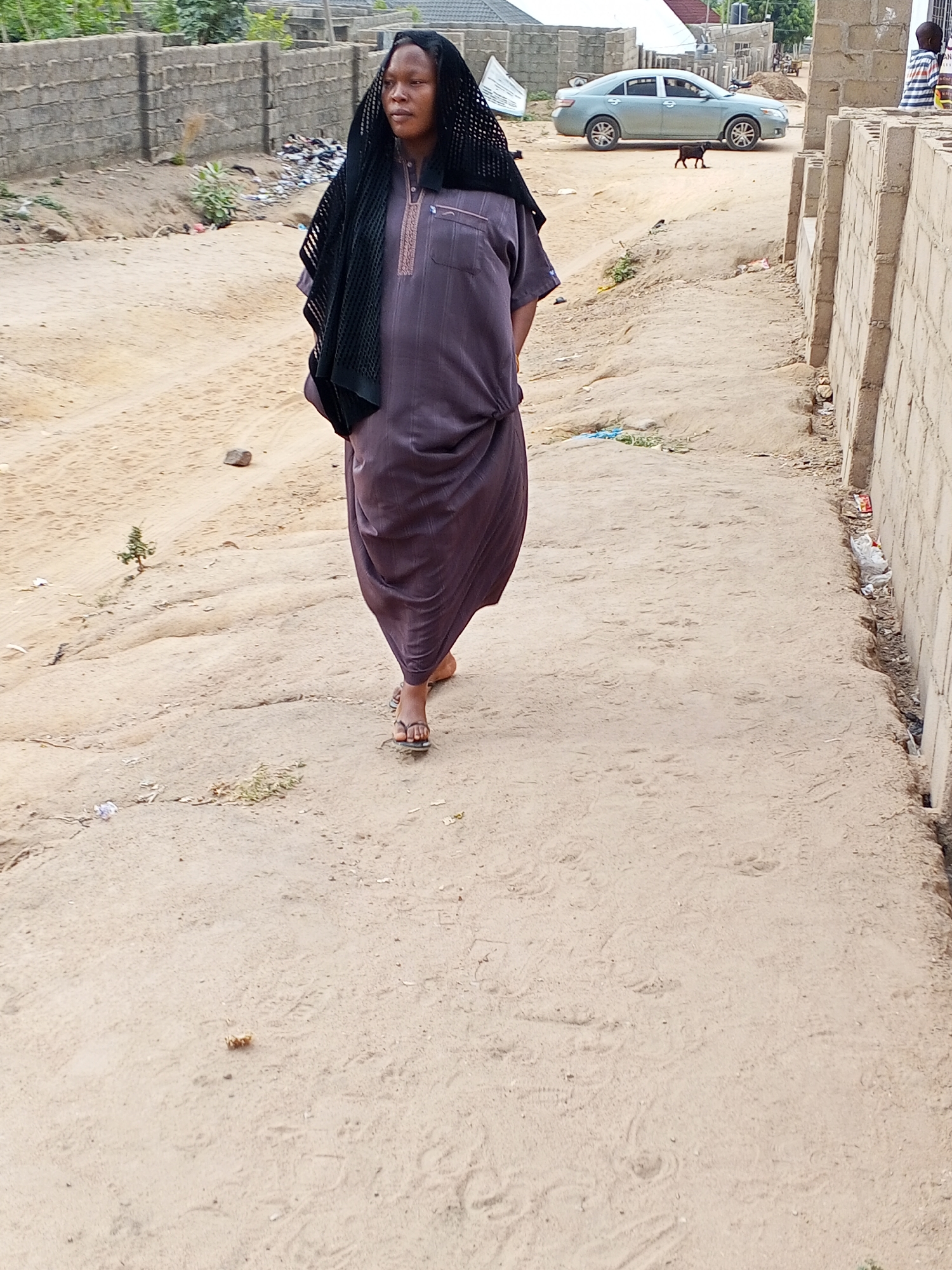The image shows a full-length view of a person walking on an unpaved, sandy road in Adonkolo, Nigeria, during daylight hours. The individual, who appears to be female, is positioned centrally and walks towards the viewer's left. She wears a long, dark purple or grey robe-like garment with light-colored embroidery around the neck, and a black, loosely-knit head covering that drapes over her shoulders. She is barefoot, wearing dark-colored sandals. Her expression is neutral, and she gazes straight ahead.
The immediate setting is a wide, sandy path marked with footprints and some small debris. To the left, a low concrete block wall runs along the path, with sparse green vegetation and some discarded items behind it. To the right, another similar concrete block wall defines the edge of the path. Further in the background, a light blue sedan car is parked on the sandy ground. A small, dark-colored animal, resembling a goat or small dog, stands near the car. Several buildings, constructed with concrete blocks, are visible further in the distance under a bright sky, suggesting clear weather. The scene depicts a common street or pathway in a semi-developed residential area. Indistinct text can be seen on the far right wall.