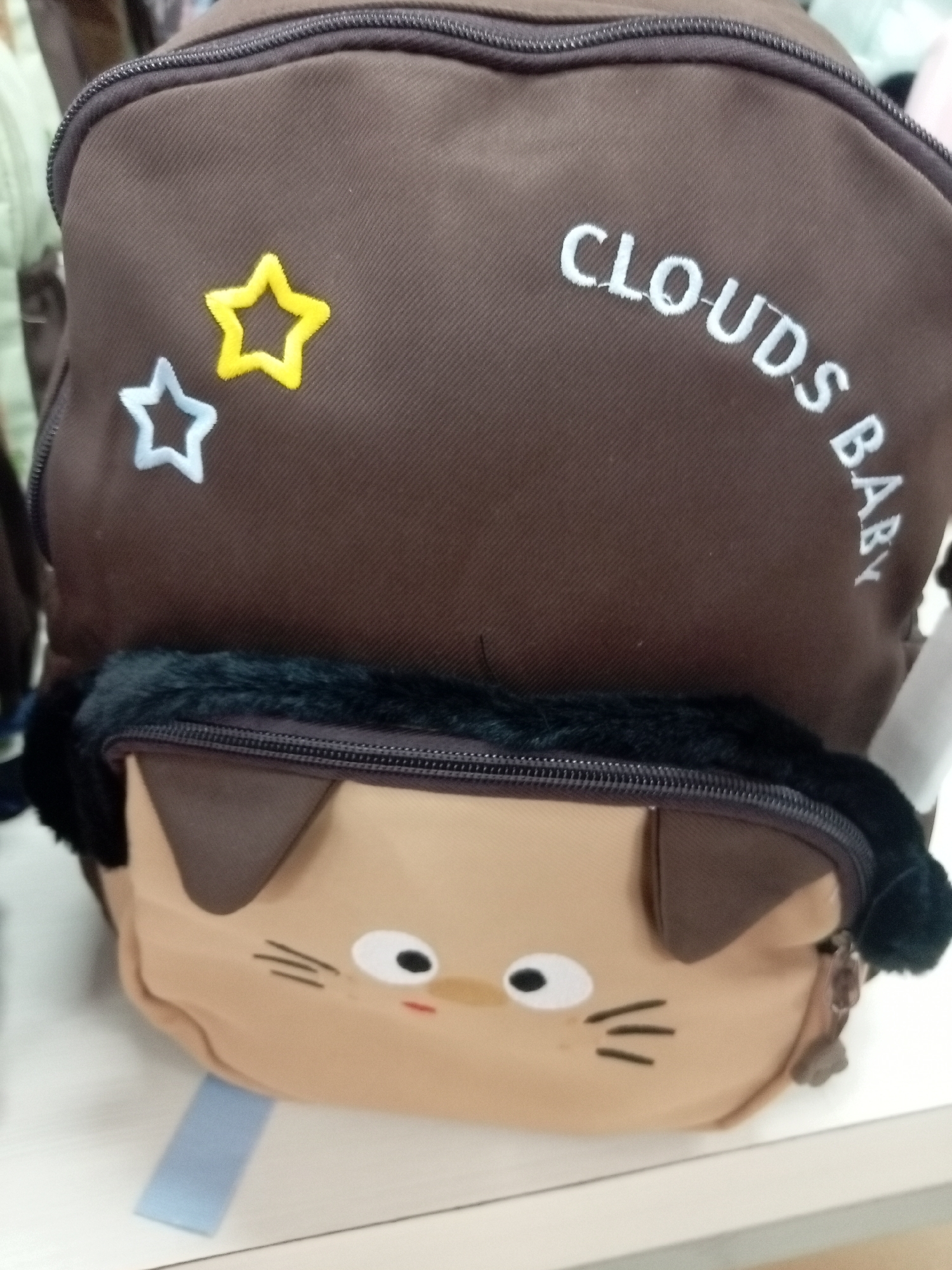 The image shows a close-up view of a brown backpack with a cartoon cat face design on the front pocket. The backpack has a fluffy black trim around the top of the pocket. There are two embroidered stars on the main body of the backpack: one yellow and one light blue. White text is embroidered on the backpack, appearing to read "CLOUDS BABY". The backpack is placed on a white surface, possibly a shelf or table. The background is blurred but appears to be indoors, with some hints of other objects or bags. There is no identifiable location in Batam, Indonesia, or any people visible in the image.