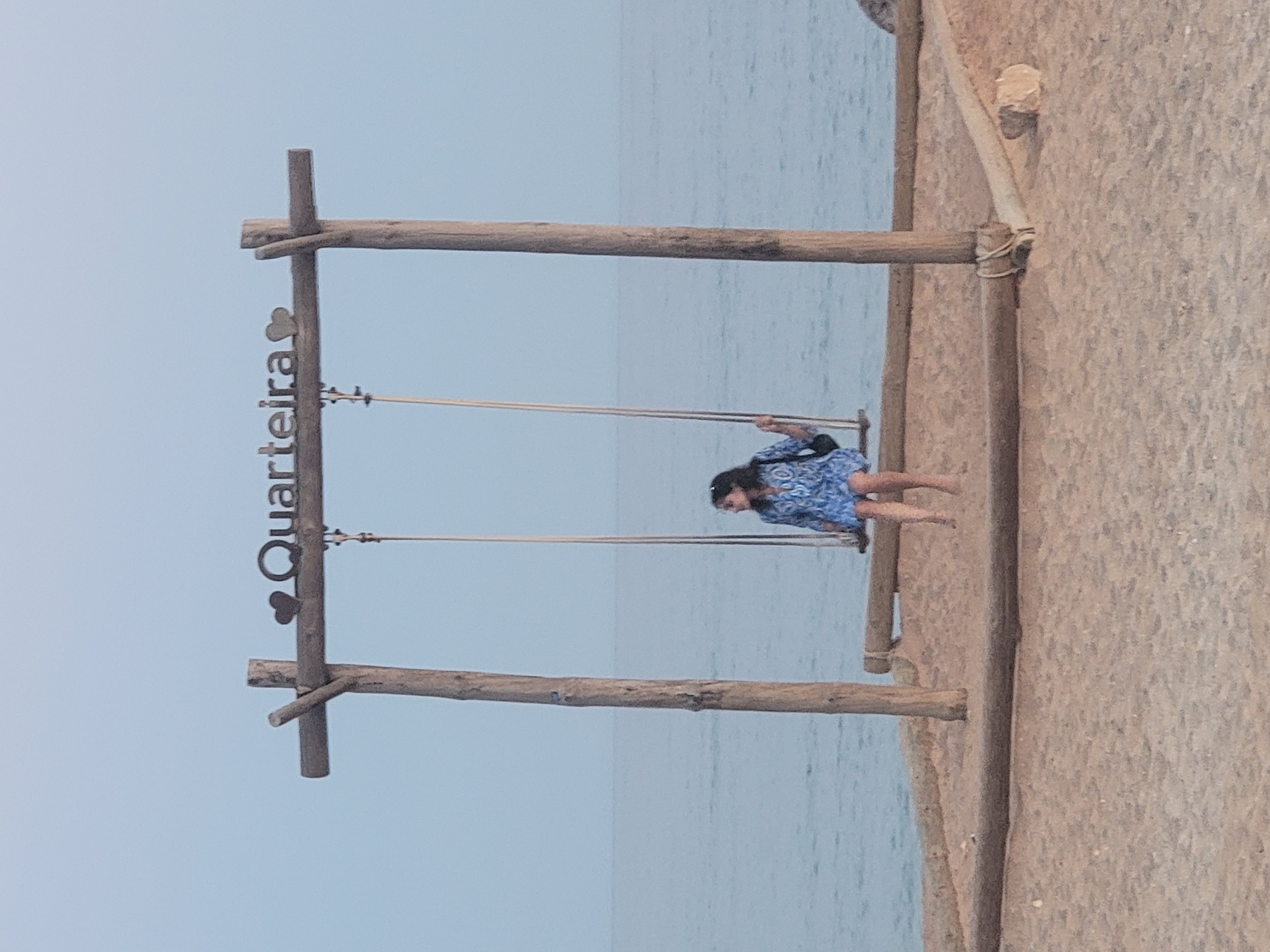 A young woman sits on a swing by the sea in Quarteira, Portugal. The swing is part of a wooden structure with the word "Quarteira" inscribed above it. The woman is wearing a blue and white patterned dress and has a black bag on her shoulder. The sea is calm and the sky is clear. The scene is set on a sandy beach.