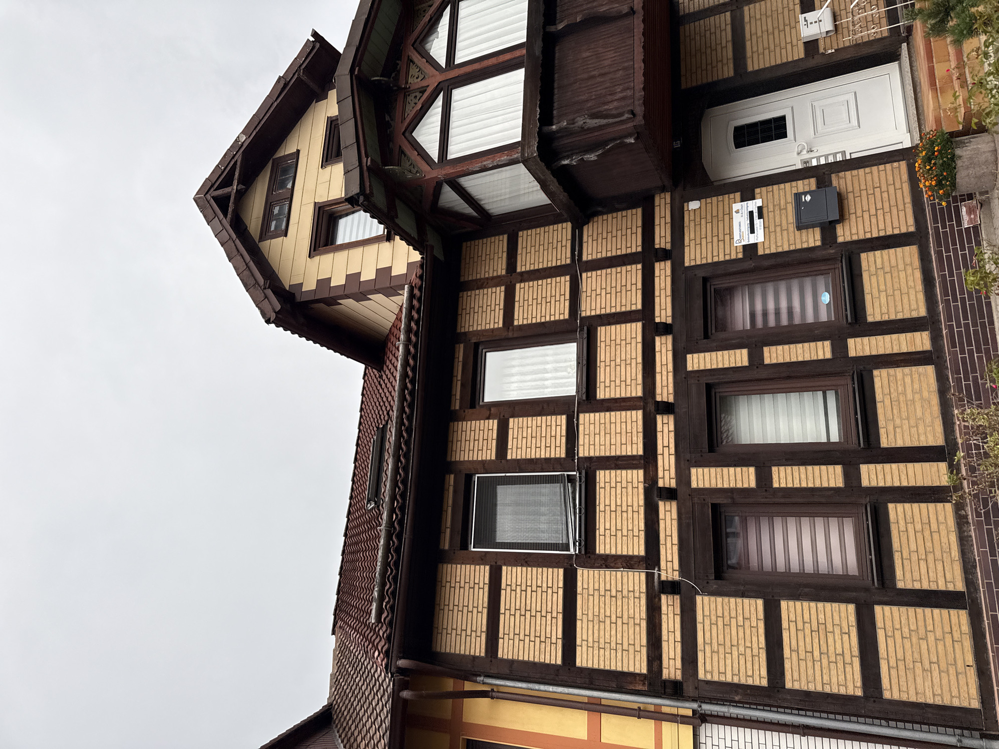 The image shows a section of a multi-story building with a distinct half-timbered architectural style. The facade features a light yellow brick pattern interspersed with dark brown timber framing. Several windows are visible, some with blinds and others with white curtains. A prominent dark brown bay window with decorative trim juts out on the upper level. A white front door with a small glass pane is located on the ground floor. A mailbox and a small sign are mounted on the wall next to the door. The roof is covered with red ceramic tiles. The sky is overcast. The building is located at Bismarckstrasse 13 in Bad Sachsa, Germany, as indicated by the sign next to the door. There are no people or animals visible in the image. The scene is outdoors, with some low-lying plants in a planter at the bottom right of the image.