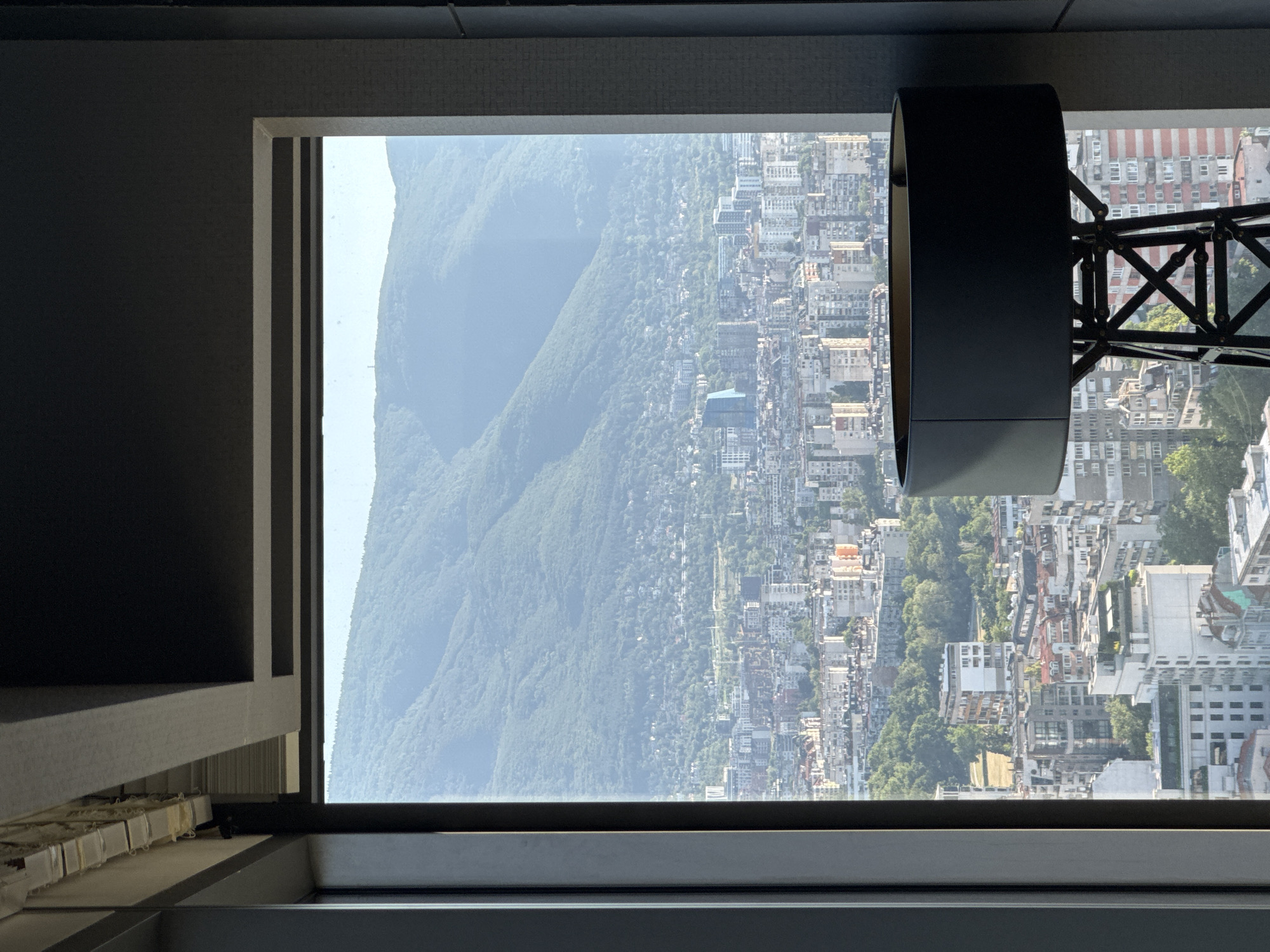 The image shows a view of Sofia, Bulgaria, seen through a large window. In the foreground, a black lamp with a cylindrical shade stands to the right. The lamp has a complex, industrial-style base made of dark metal with crisscrossing supports.
Beyond the lamp, the window frames a panoramic view of the city. Numerous buildings of varying heights and architectural styles are densely packed together, with some older, lower-rise structures interspersed with more modern, taller apartment buildings. The buildings are primarily residential, with facades in shades of white, beige, and some with orange accents. Green trees are visible in pockets throughout the urban landscape, suggesting parks or residential gardens.
In the background, a large, verdant mountain range dominates the horizon. The mountains are covered in lush green trees, indicating a forested slope. The sky is clear and bright, suggesting it is daytime, likely morning or afternoon given the soft light. The overall impression is of a city nestled against a natural backdrop. The window frame itself appears to be part of a modern building, possibly a hotel or a high-rise apartment complex, given the elevated perspective.