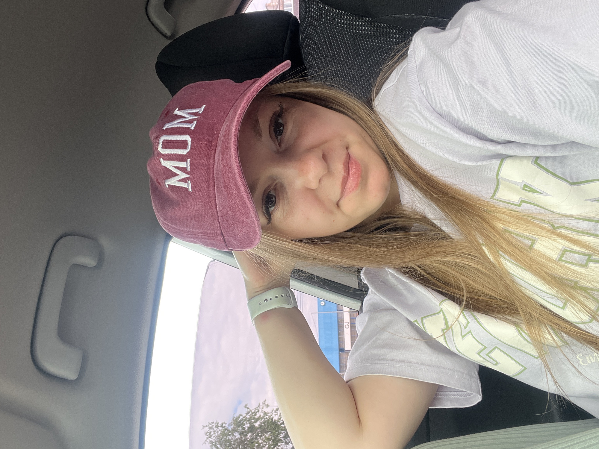 A young woman with blonde hair is sitting in the passenger seat of a car, wearing a maroon baseball cap with "MOM" embroidered on it in white lettering. She is also wearing a light purple t-shirt with green lettering. Her left arm is resting on the car's headrest, and she is looking at the camera with a slight smile. Through the car window, a cloudy sky and some trees are visible. A building with blue accents and the letters "GJ" can be seen in the distance. The car interior includes a grey grab handle and a dark grey car seat. The image was taken on a cloudy day.