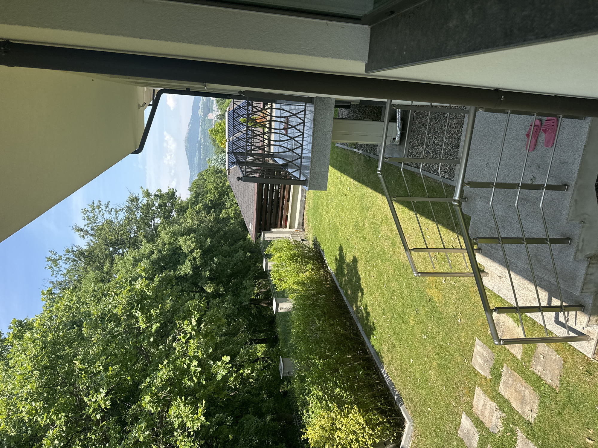 This image depicts an outdoor residential scene in Rezbartsi, Bulgaria, under bright daylight with a clear blue sky. The viewpoint is from an elevated position, looking down and across a private garden area.
In the immediate foreground, to the right, is a light-colored exterior wall of a building with a dark vertical downspout. A dark, flat sill or ledge extends from the wall. A set of outdoor stairs, constructed from light gray granular stone, descends from the viewer's perspective. These stairs feature a brushed stainless steel handrail with multiple horizontal bars. Two pink plastic slip-on sandals are resting on the upper steps. Below the stairs, a dark gravel-covered ground area is partially visible.
To the left of the stairs and extending into the midground is a well-maintained green lawn. A pathway of irregular rectangular stepping stones is laid across the lawn. Along the far edge of the lawn, a dense, manicured hedge of green bushes runs horizontally, bordered by a concrete curb. Behind the hedge, a wire fence is visible, supported by several concrete posts with square caps.
Further into the midground, beyond the hedge and fence, a small, dark-roofed building, possibly a gazebo or shed, is partially visible. Adjacent to this structure, a cantilevered balcony or terrace extends outward, featuring an ornate dark metal railing with a geometric pattern and a reflective dark floor. Above and behind this balcony, solar thermal collectors (likely evacuated tubes) are visible, catching the sunlight.
The background features a dense expanse of lush green trees, indicating a forested area. Above the treeline, under a partly cloudy blue sky, distant mountains or hills are visible, with one prominent peak. A glimpse of urban development, including buildings, can be seen in the valley below the distant mountains, suggesting a nearby city or town. The sunlight casts distinct shadows, notably from the railings onto the stairs and from the building onto the lawn. No people are present in the scene.