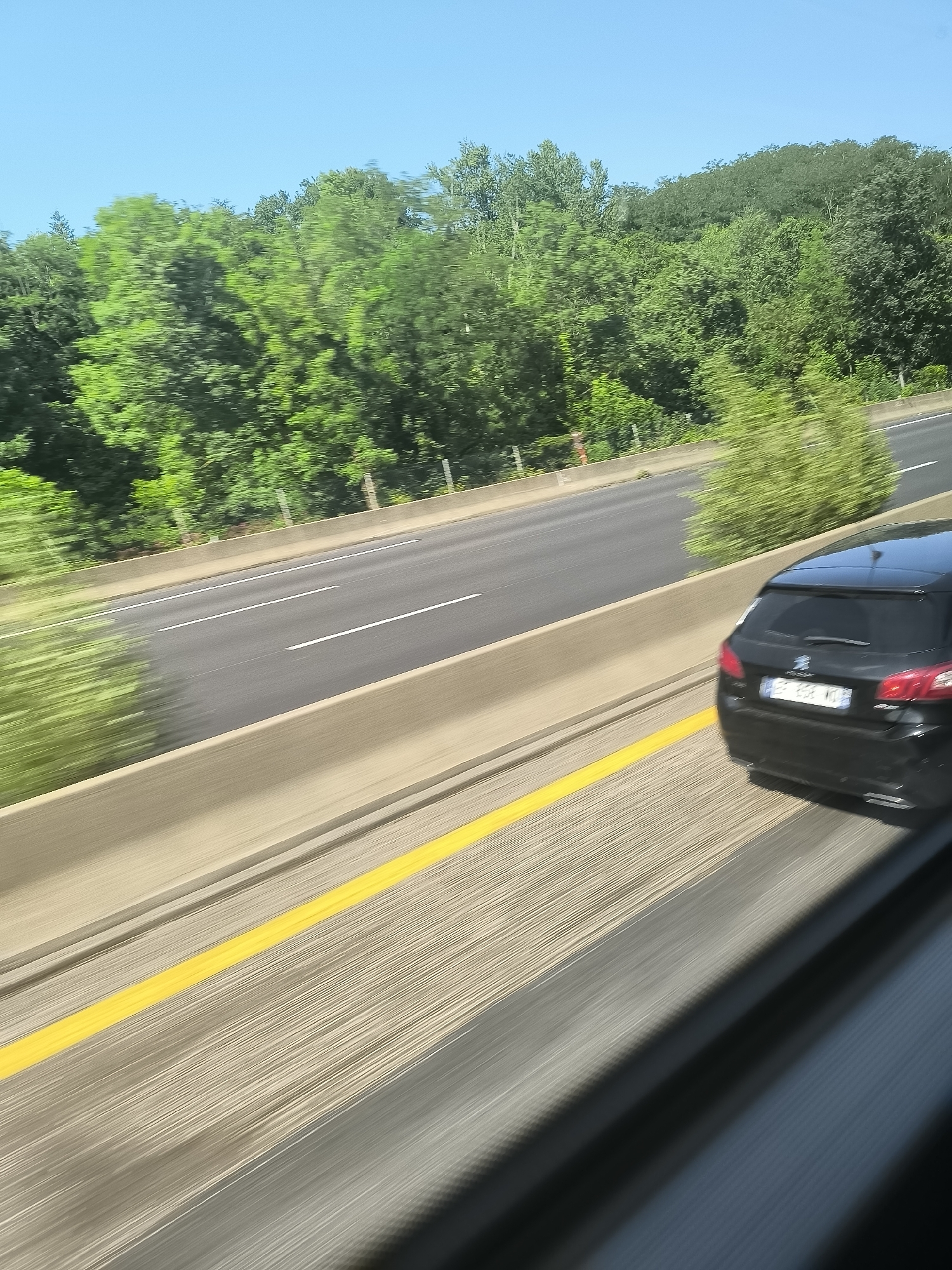 A black Peugeot 308 station wagon is traveling on a highway bordered by a concrete barrier, to the right. The highway has multiple lanes separated by white lines and a solid yellow line along the barrier. A green treeline is in the background under a clear blue sky. The photograph appears to have been taken through a window while in motion. The image contains motion blur. The license plate on the Peugeot is blurred, but partially visible with the characters "EB-258-VK". The location is likely near L'Horme, France, given the license plate style and the presence of signs in the background that are consistent with French roads.