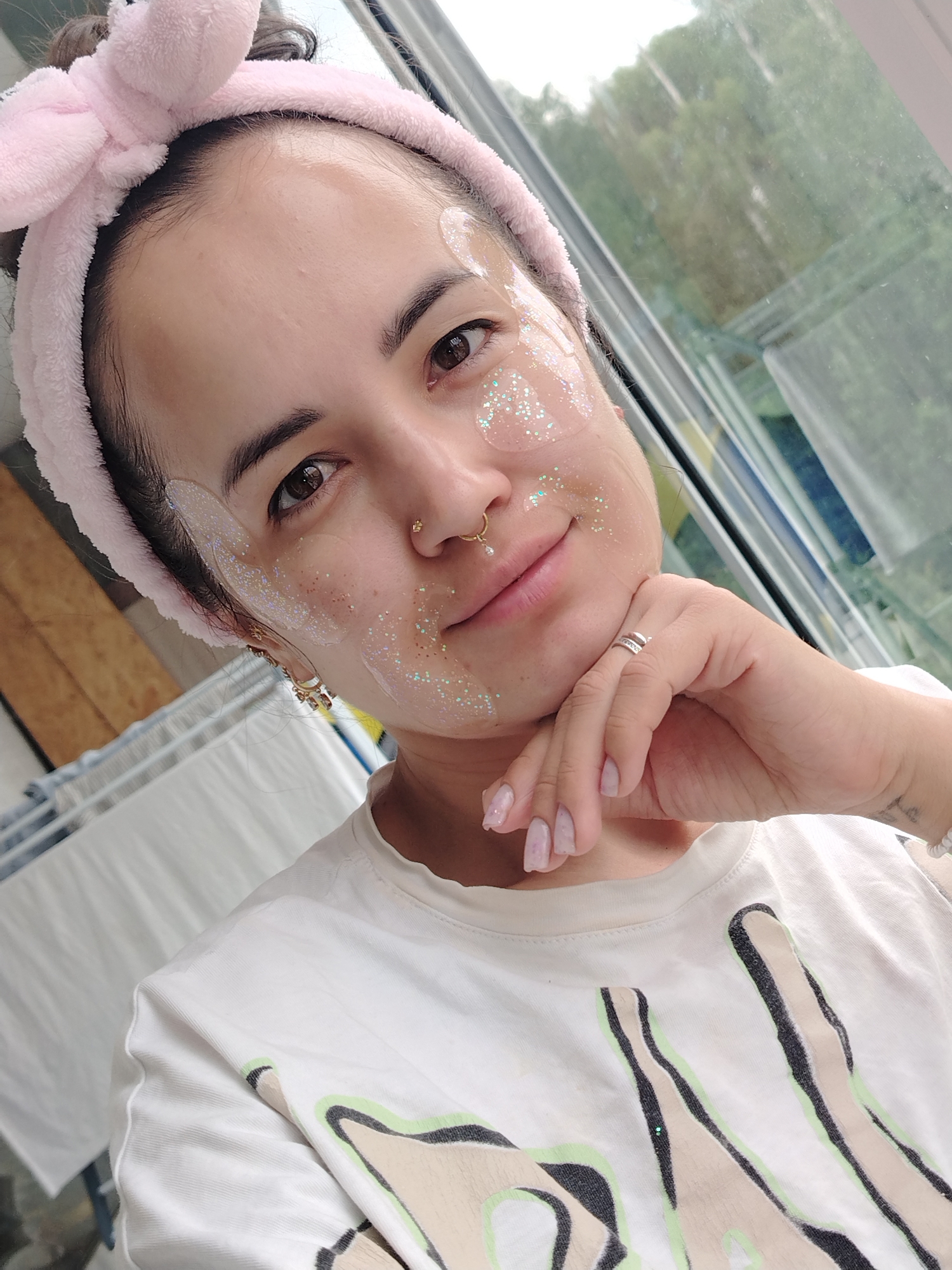 A person with fair skin, dark eyes, and dark hair pulled back by a soft pink bow headband is depicted in a close-up selfie. They are wearing clear, glittery face patches on their cheeks, forehead, and jawline, suggesting a beauty or self-care routine. Two golden nose rings, one a hoop and one a stud, are visible, along with an earring on their left ear. Their chin rests on their right hand, which displays a silver ring with small stones on the ring finger, and fingernails painted with a light, iridescent polish.
The person has a gentle smile, looking directly at the camera. They are wearing a white t-shirt featuring an abstract graphic design in beige, black, and light green colors. In the background, a large window reveals a view of green trees and foliage, indicating an outdoor, natural setting. To the left, a white clothing drying rack is partially visible. The scene appears to be an indoor setting, likely a home, with natural light streaming through the window, suggesting it is daytime. The location is in Noviy, Russia. No visible text is present on the clothing or in the immediate surroundings.