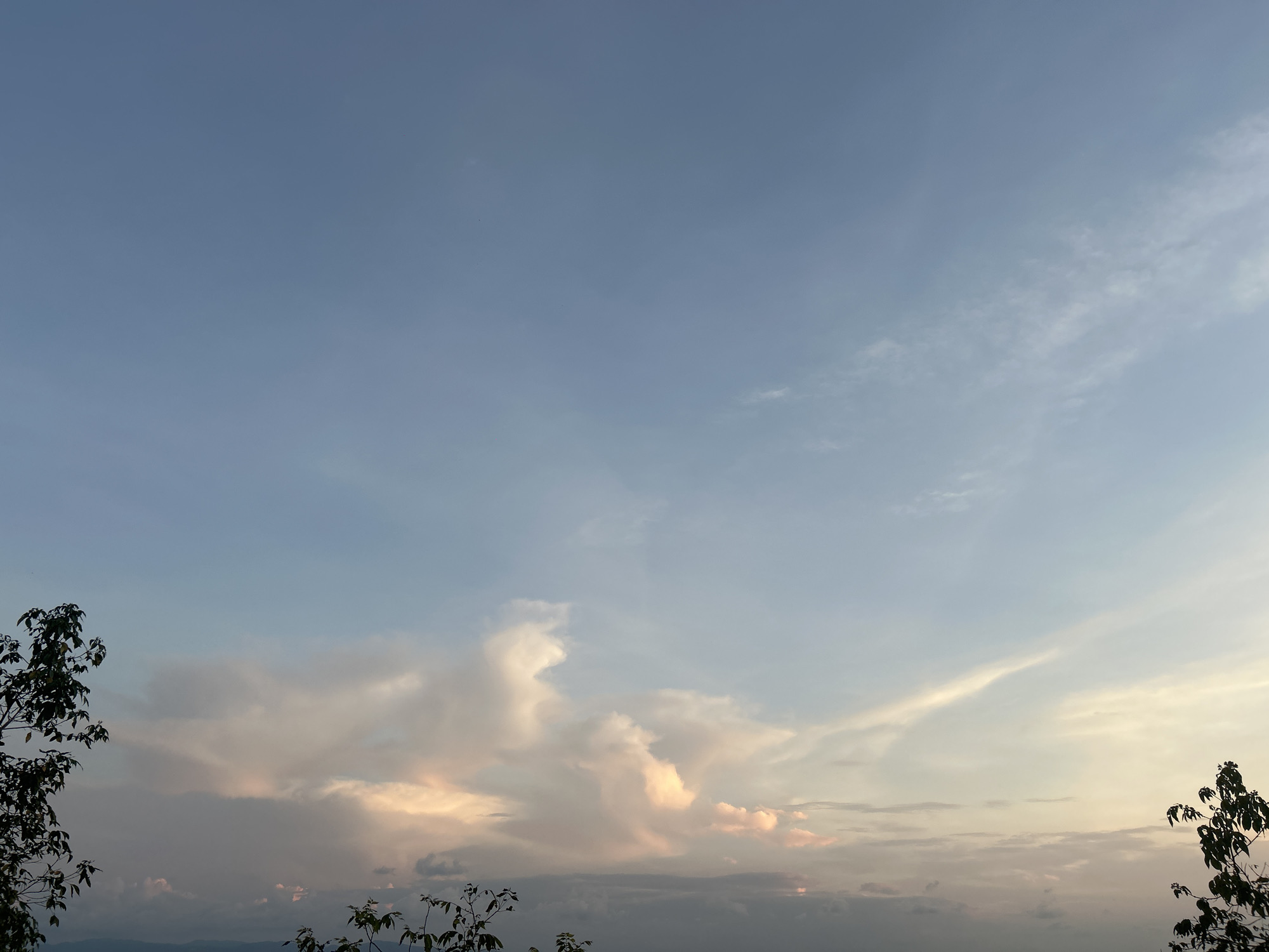 The image shows a sky in Baan Hin Kong, Thailand. The sky is a gradient of blue and pale white, with the blue more prominent in the upper portion. There are clouds in the lower-middle of the image, tinged with a golden hue, suggesting sunset or sunrise. Silhouetted treetops are visible along the bottom of the frame and on either side, obscuring the horizon.