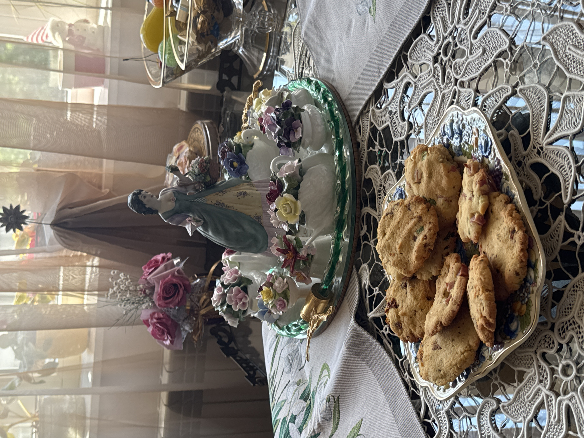 A still life photograph shows a display of cookies on a decorative plate, a porcelain figurine, flowers, and other items arranged on a table. The cookies are on a blue and white patterned plate, while the figurine depicts a woman in a dress, surrounded by floral embellishments on a decorative base. A bouquet of roses is in the background near the figurine. The objects are set against a window with sheer curtains. The location is impossible to determine based on the contents of the image.