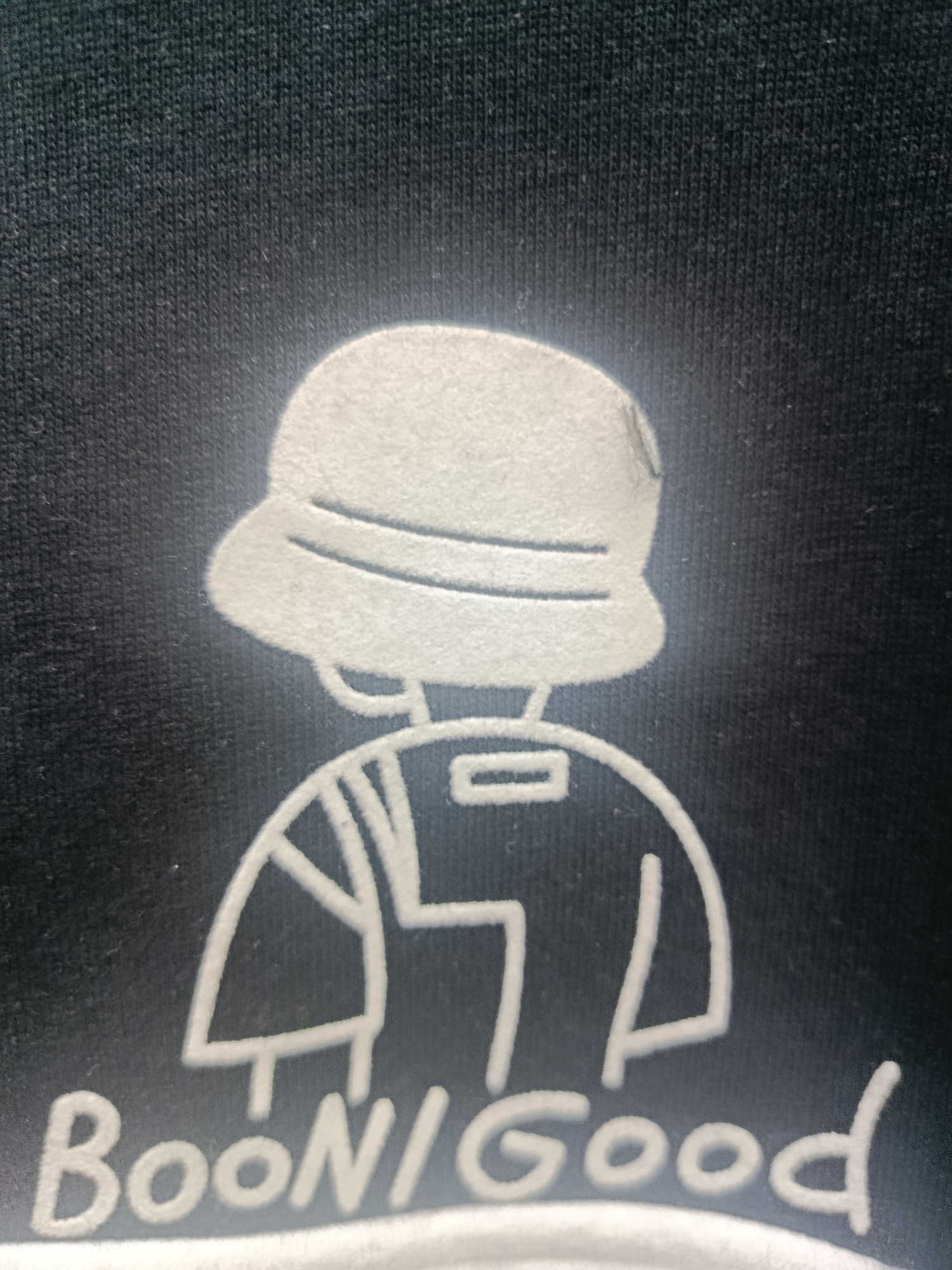 The image shows a close-up of a dark fabric, possibly a t-shirt, with a white graphic printed on it. The graphic depicts a stylized drawing of a person wearing a bucket hat and a shirt with some sort of design on it. Below the figure, the text "BooN/Good" is written in a bold, white font. The image is taken from a low angle, and the lighting creates a slight halo effect around the graphic. The fabric has a visible texture of vertical knit lines. The scene is indoors, and there are no other objects or people visible. The location of Sertolovo, Russia is not depicted in this image.