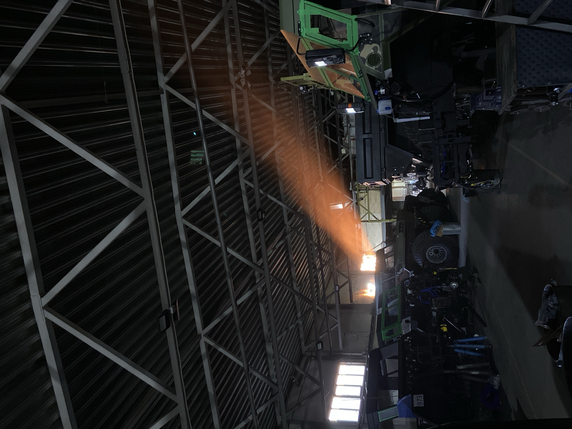 The image shows the interior of a large industrial building, possibly a factory or warehouse, in Krasnoyarsk, Russia. The roof structure is visible, constructed of metal beams and corrugated sheeting. Light streams in through windows, creating a strong beam of light. Machinery, possibly construction or industrial equipment, is present. No people are directly visible, but the presence of the machinery implies human activity. A number "973" is seen on a blue object near the base of one piece of machinery.