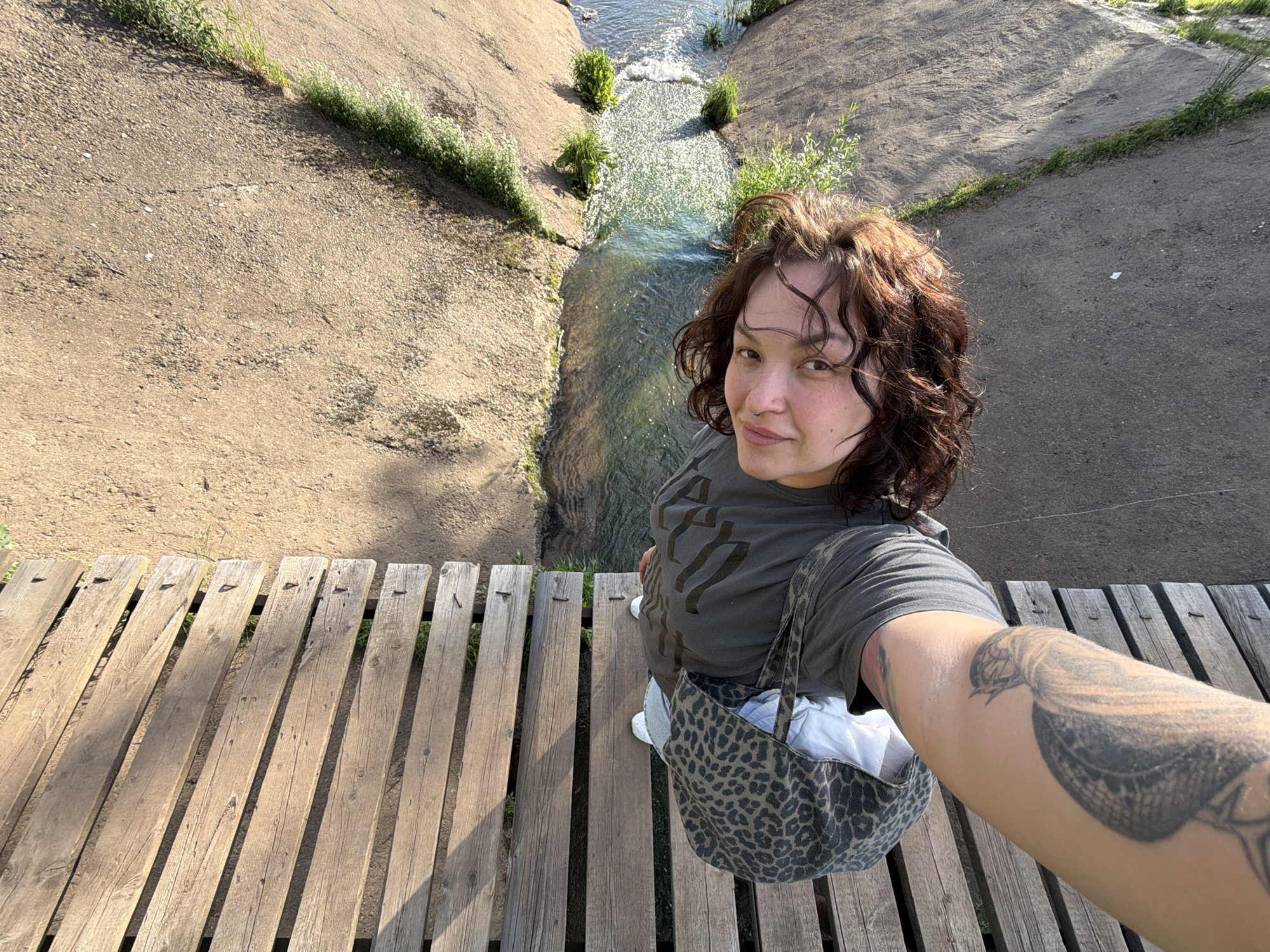 The image shows a young person with curly brown hair and fair skin, taking a selfie from a high vantage point. They are wearing a dark t-shirt and leopard print shorts, with white sneakers. The person is standing on a wooden walkway that appears to be part of a viewing platform or bridge. Below the walkway is a body of water, possibly a river or stream, with rocky banks and some vegetation. The overall scene suggests an outdoor natural setting. The orientation of the image indicates the photo was taken in portrait mode and then rotated. The person's arm is visible in the foreground, showing a tattoo on their upper arm. The background consists of sandy ground and some green grass. There is no visible indication of the city of Chaikovsky, Russia.