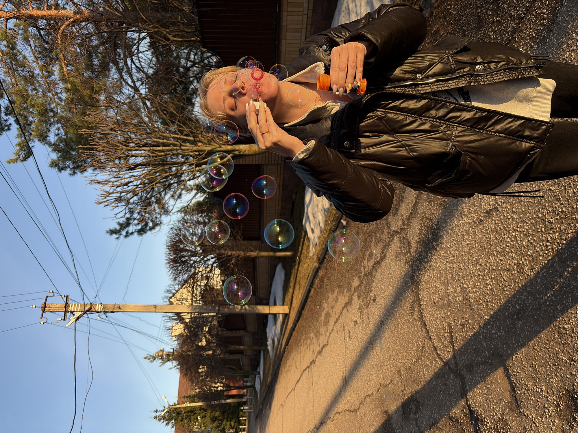 A young woman stands on a paved street, blowing bubbles. She wears a black puffer jacket, a white hooded sweatshirt, and black leggings. In her right hand, she holds an orange and white bubble solution container, while her left hand holds a bubble wand. Multiple iridescent bubbles float in the air around her. The location appears to be a residential area in Прикамский, Russia, evident by the presence of utility poles with wires, a sidewalk, bare trees, and houses in the background. The scene is bathed in warm, late afternoon sunlight, casting shadows on the street.