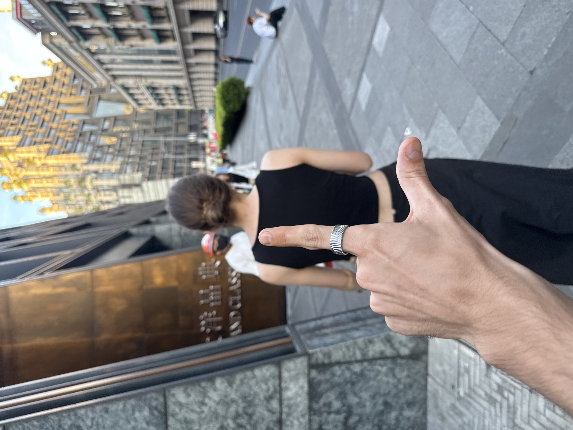 The image, oriented sideways, captures an urban scene from a first-person perspective. In the immediate foreground, a right hand is prominently displayed on the lower right, making a "thumbs up" gesture with the index finger extended and adorned with a distinctive silver-colored ring featuring a wavy or ridged texture.
In the mid-ground, slightly to the left and further back, the back of a person with dark hair (possibly in a bun) is visible, wearing a black sleeveless top and dark pants or a skirt. They are facing away from the camera, looking towards the background.
The background reveals a bustling city environment. Tall, modern buildings with varied architectural styles, including glass facades and light-colored exteriors, dominate the left side of the frame. One prominent building features a large, reflective golden-brown sign at its base. On this sign, partial Chinese characters are visible alongside the English words "AND CLASS." The ground is paved with large gray square tiles, suggesting a plaza or wide sidewalk. Further in the distance, blurred figures of other people are scattered, including one individual sitting on the ground wearing a white shirt and dark pants. Green shrubs or hedges line parts of the path.
The lighting suggests late afternoon or early evening, with a warm, golden glow illuminating parts of the buildings. The sky is bright but not directly visible. The overall scene depicts a casual public setting in Putuo District, China, conveying a sense of urban life and possibly a positive sentiment through the "thumbs up" gesture.