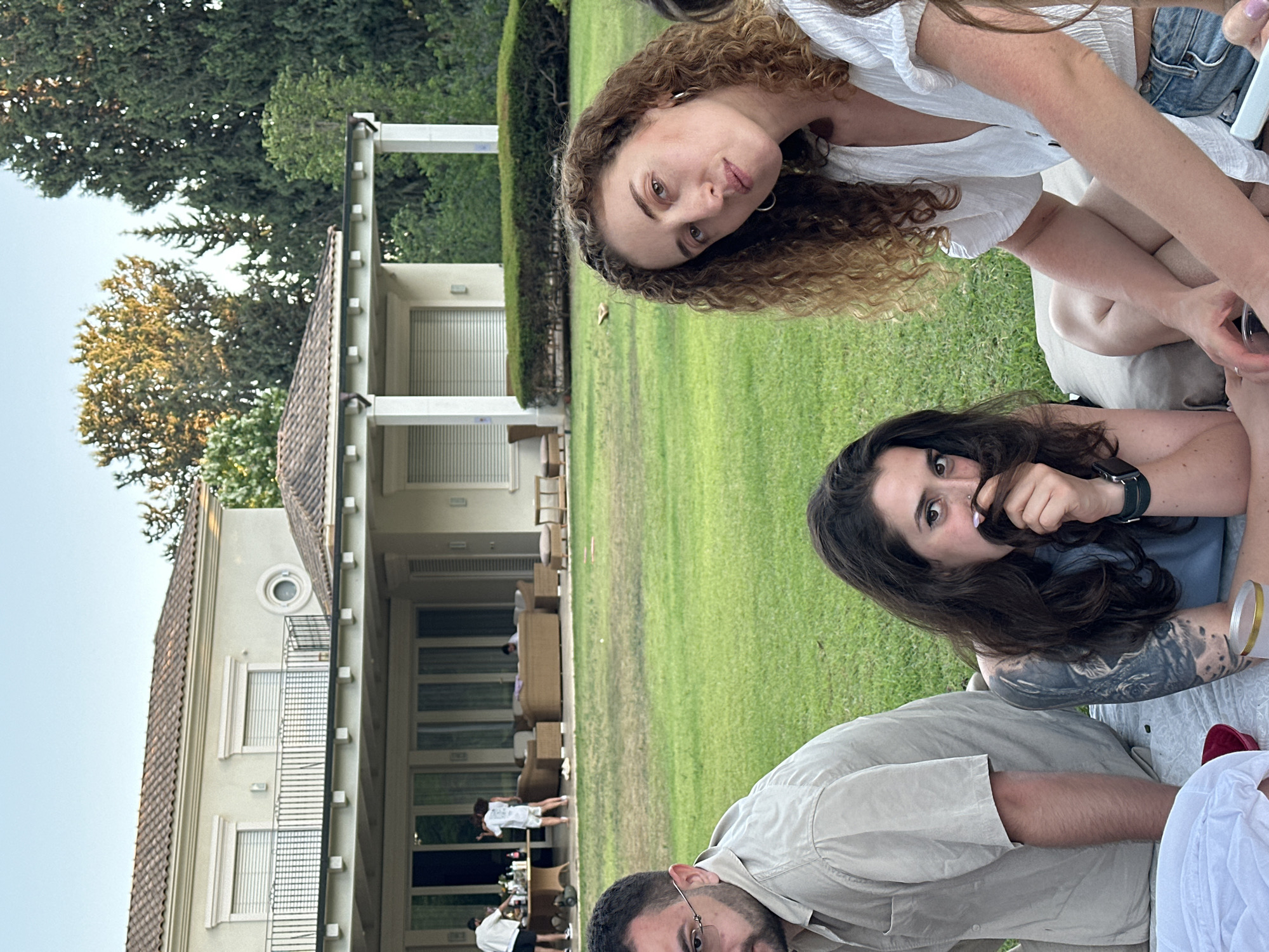 In Savyon, Israel, an outdoor gathering unfolds on a manicured lawn in front of a two-story villa with a tiled roof and expansive windows. Four individuals are seated on a light-colored blanket, facing the camera.
To the left, a man with dark hair and glasses is positioned. He wears a light-colored, short-sleeved button-down shirt. Next to him, a woman with long dark hair, partially obscuring her face, is visible. She has tattoos on her left arm and wears a dark-colored top. She is holding a small object to her mouth with her hair.
In the center-right, a woman with curly auburn hair is seated, looking directly at the camera. She wears a white, short-sleeved top and denim shorts. Her hands are clasped in her lap. To her right, another individual is partially visible, wearing a white top.
In the background, several people are standing near a table laden with bottles and glasses, suggesting a party or event. Another group is seated on outdoor furniture on a covered patio. The environment is characterized by lush green grass, mature trees, and clear sky.