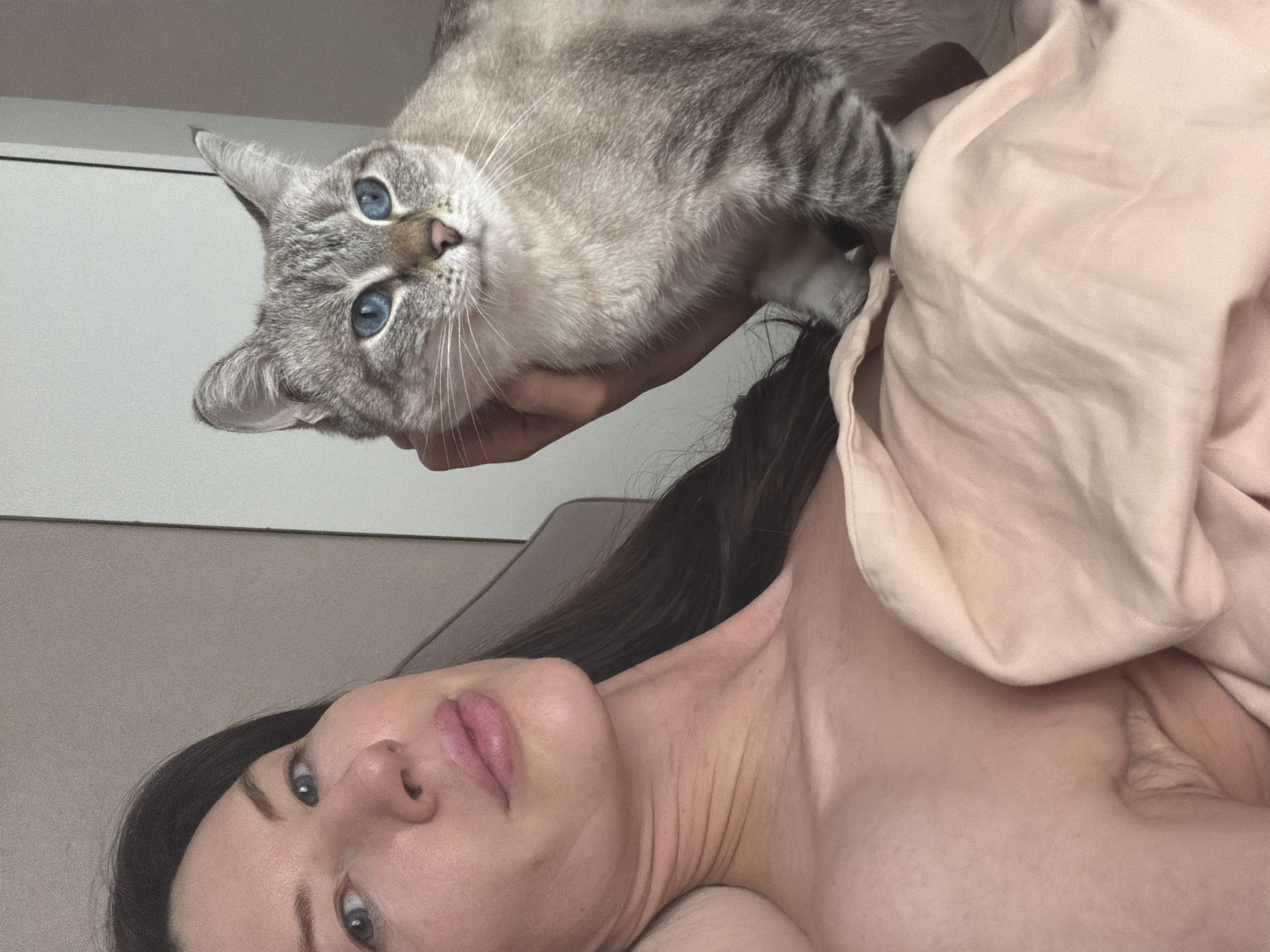 The image presents a close-up, rotated view of a woman and a cat in an indoor setting, likely a bedroom, with the provided context indicating Perm, Russia.
The central subjects are a woman and a light gray tabby cat. The woman is lying down, partially covered by a light pink or beige blanket. Her face, showing dark hair, blue-gray eyes, and soft pink lips, is visible, and she looks directly towards the camera with a calm expression. Her neck and shoulder are also visible.
Above her, a hand (presumably hers) holds the cat, positioning its head prominently in the frame. The cat is a striking feature with its bright blue eyes, pink nose, and long white whiskers. Its light gray fur displays distinct darker tabby stripes, and it too is looking towards the viewer with a curious and direct gaze.
The background is simple, consisting of a light-colored wall or headboard. The overall lighting is soft and even, suggesting a well-lit indoor environment. The scene conveys a moment of quiet interaction and affection between the woman and her cat. No text is visible in the image.