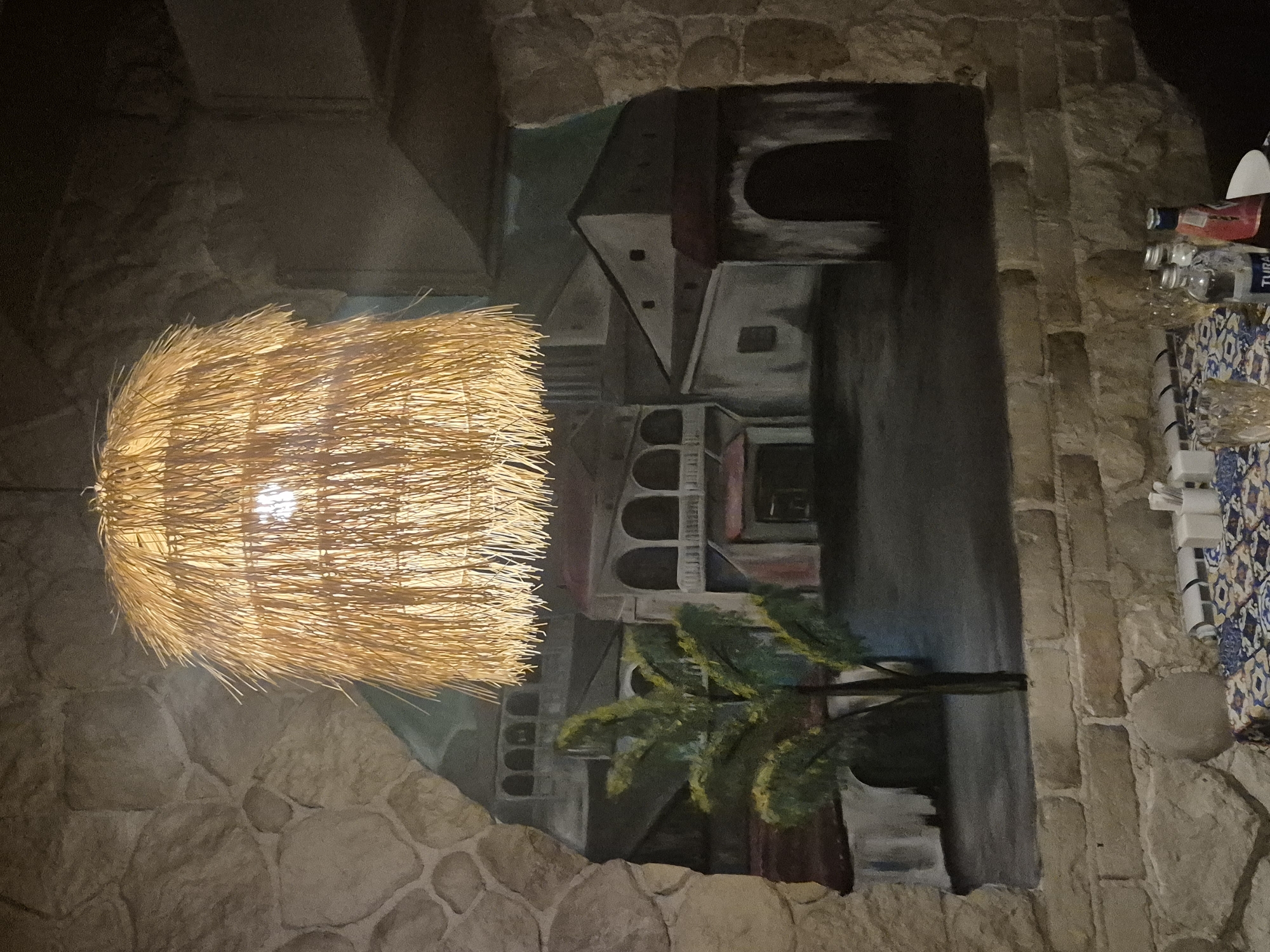 The image depicts an indoor scene, likely a restaurant or cafe, with a distinctly rustic and artistic ambiance. The most prominent feature is a large, woven straw lampshade hanging from the ceiling, casting a warm, diffused light. Behind the lampshade, a mural is painted on the wall, creating the illusion of a European town or village with traditional architecture, including arched windows and balconies, and a lone palm tree. The walls themselves are constructed of rough, uneven stone and brickwork, enhancing the textured and aged feel of the space. In the foreground, a table is set with a patterned tablecloth, glassware, and several bottles, suggesting a dining area. The overall impression is one of a thoughtfully designed space that blends natural materials with artistic expression, potentially aiming for a cozy and unique dining experience. There are no people visible in the image. The location within the city of Kokshetau, Kazakhstan, cannot be determined from the visual information provided in the image.