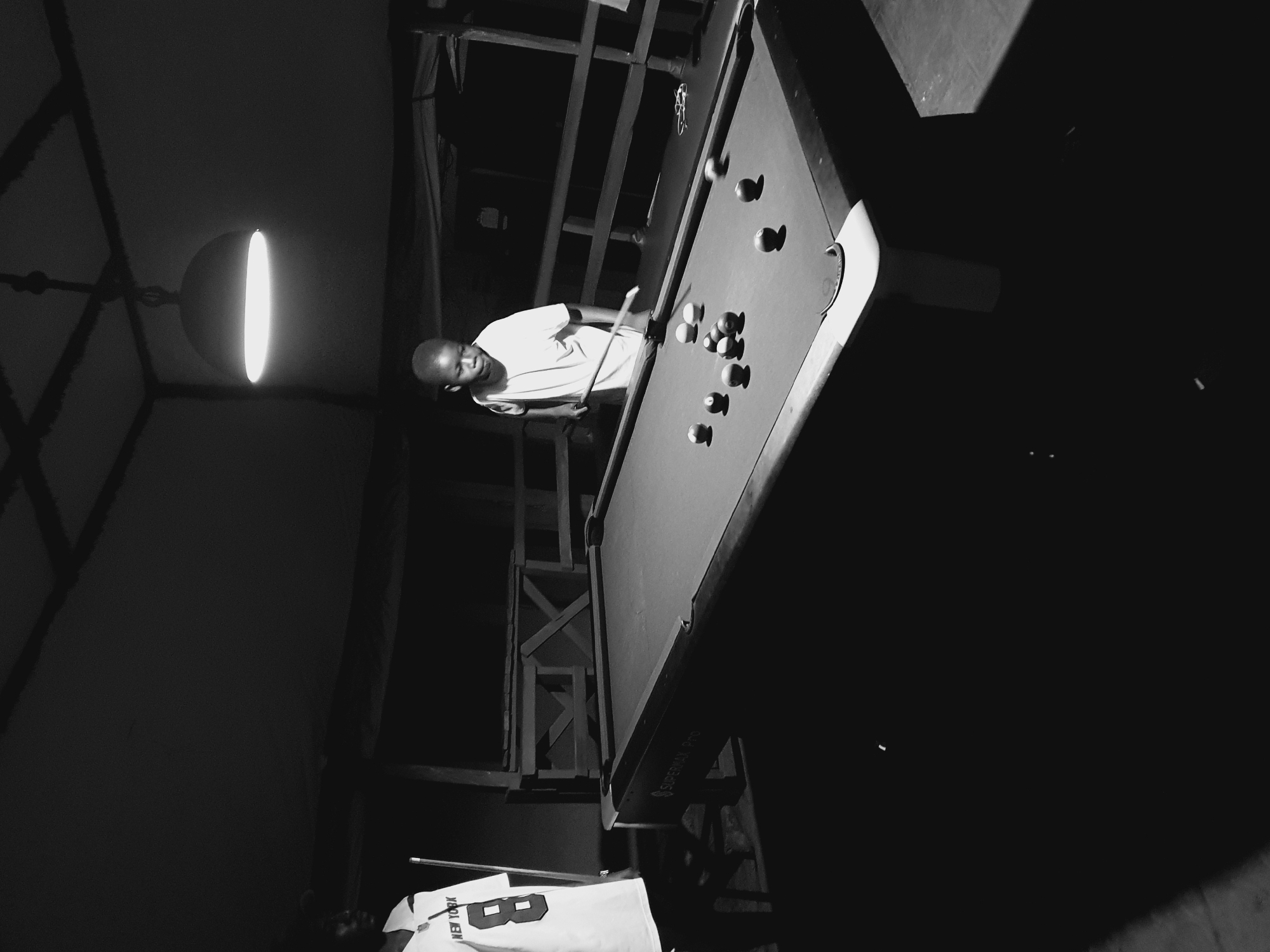 A black and white indoor shot depicts a man playing pool. He is wearing a light-colored shirt. A pool table with balls is visible, illuminated by an overhead light. A partially obscured shirt with "NEW YORK" printed on it lies to the left. The location in Lokoja, Nigeria cannot be determined.