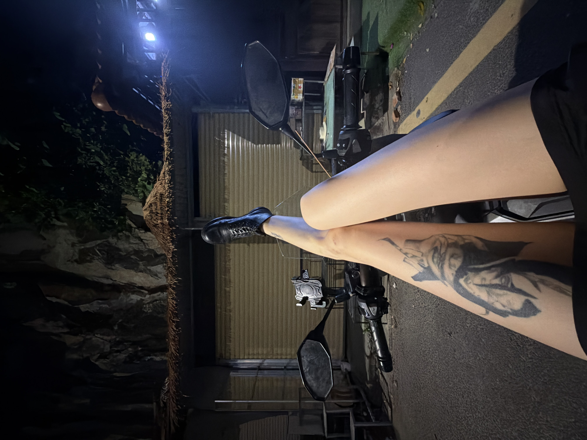 The image shows a person sitting on a motorbike at night. The person's legs are visible, with a black boot on one foot and a tattoo on the other leg. The motorbike is parked on a street with a yellow line. In the background, there's a building with a closed rolling shutter. To the left, there's a wall or rock face with some vegetation and a light fixture. The location is likely in Đà Nẵng, Vietnam.