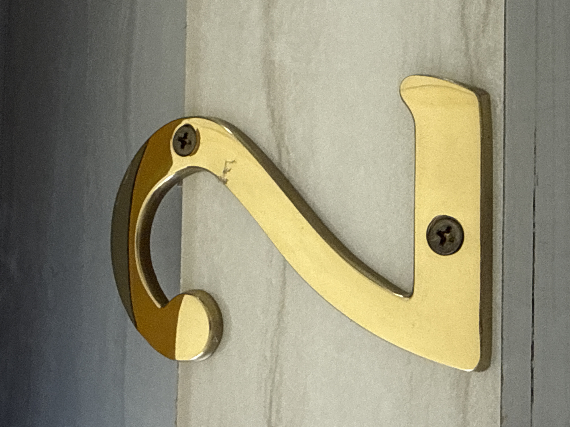 A close-up, eye-level shot features a gold-colored numeral "2" mounted on a light-colored, textured surface. The numeral is made of metal and has a smooth, polished finish, reflecting light and showing some minor scuffs and wear. It is secured to the surface by two dark-colored screws, one near the top curve and another near the bottom base. The background consists of a gradient of gray and blue, with subtle horizontal lines, suggesting a wall or paneling. The scene is static and lacks any human presence or activity. There is no discernible information in the image to connect it to Lhokseumawe, Indonesia, or any specific urban location.