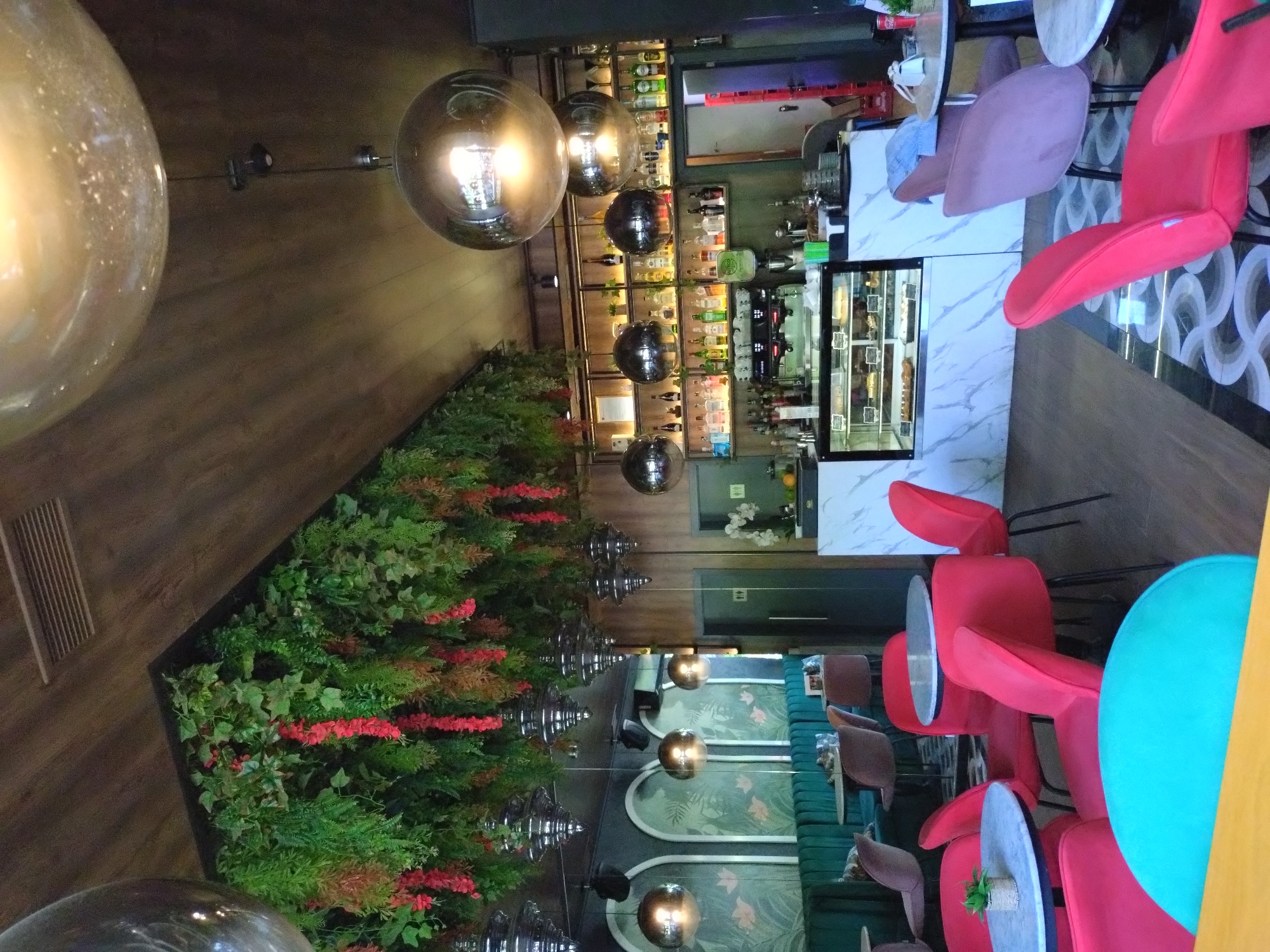 This media file depicts the interior of a modern and stylish cafe or bar, characterized by its contemporary design and inviting atmosphere. The scene is set indoors, likely at a time when there are no patrons present, possibly early morning, late evening, or a quiet period, as no people are visible.
The space is illuminated by several large, spherical pendant lights, with a prominent amber-toned one in the foreground and multiple smoky-grey, reflective globes hanging at various heights throughout the room. The flooring is a dark wooden laminate or parquet, contributing to the warm ambiance.
On the right side of the image, a well-stocked bar area features a white marble-patterned counter. Behind it, shelves display numerous bottles of spirits and other beverages. A glass display case filled with pastries or desserts is integrated into the bar counter. Adjacent to the bar, there are seating arrangements with tables and chairs. These include several red, modern-style chairs, and some lighter pink or purple chairs. A distinctive turquoise round table stands out in the foreground. Further into the background, plush dark teal or green upholstered benches provide additional seating.
A striking feature on the left is a long, raised planter acting as a "green wall," filled with lush artificial foliage and accented with vibrant red floral arrangements. This botanical element adds a fresh and lively contrast to the dark wood and metallic fixtures. A wooden slatted panel is visible on the far left wall. On the far wall near the green benches, there are decorative wall panels featuring a tropical leaf pattern. No discernible text is visible on any objects or signs within the image. The overall impression is one of a chic and comfortable establishment, likely designed for relaxation and socializing.