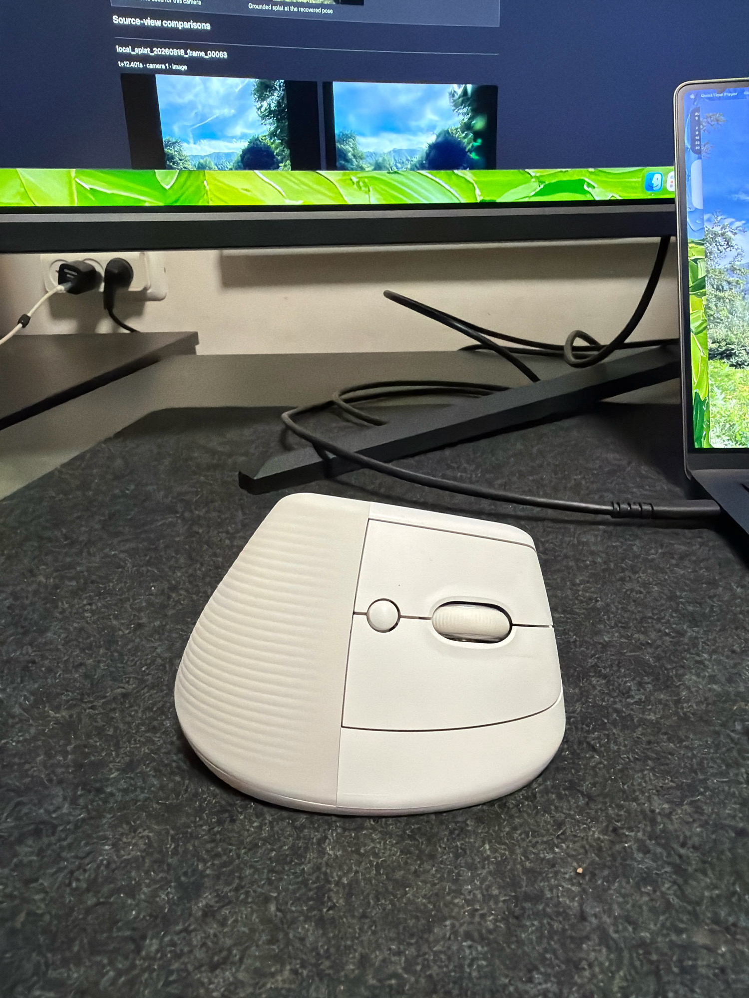 A white ergonomic mouse rests on a dark gray textured surface, likely a mousepad. The mouse has a ribbed texture on its left side and features two buttons and a scroll wheel on its top surface. In the background, a computer monitor displays a video or image with green foliage and a blue sky. To the right of the monitor, a laptop screen shows a similar outdoor scene. Power outlets with black and white plugs are visible on the wall behind the monitor. The overall scene suggests a home office or workstation setup in the city of Kafr Yasif, Israel.