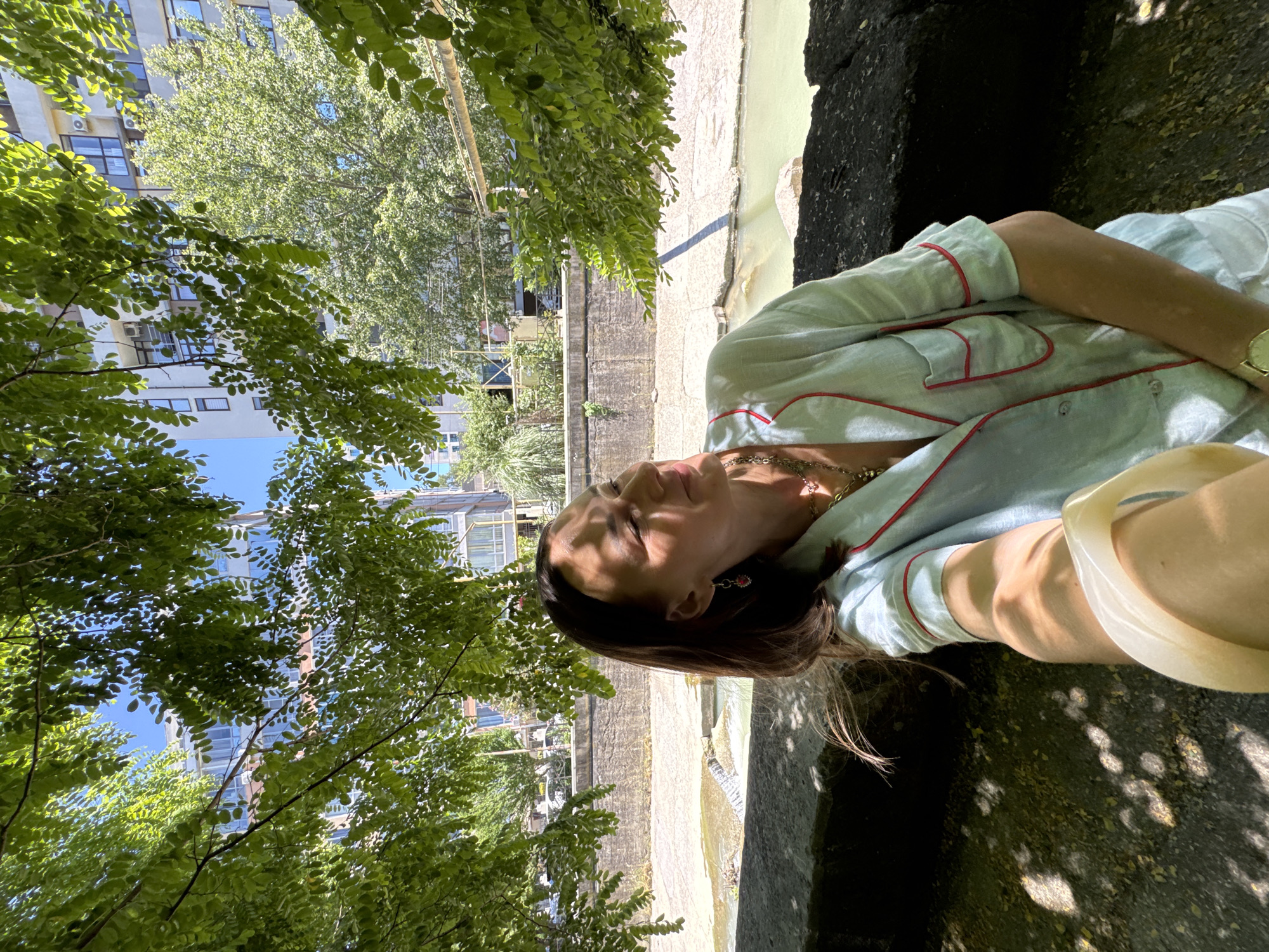 A woman is taking a selfie while sitting on a stone wall outdoors. She is wearing a light blue pajama-style top with red trim and is looking to her right, with sunlight on her face. She has on a watch and several necklaces. Behind her, a body of water flows through a channel with concrete walls. Trees with green leaves are in the foreground, and buildings and more trees are visible in the background under a clear blue sky. The image appears to be taken in Sochi, Russia.